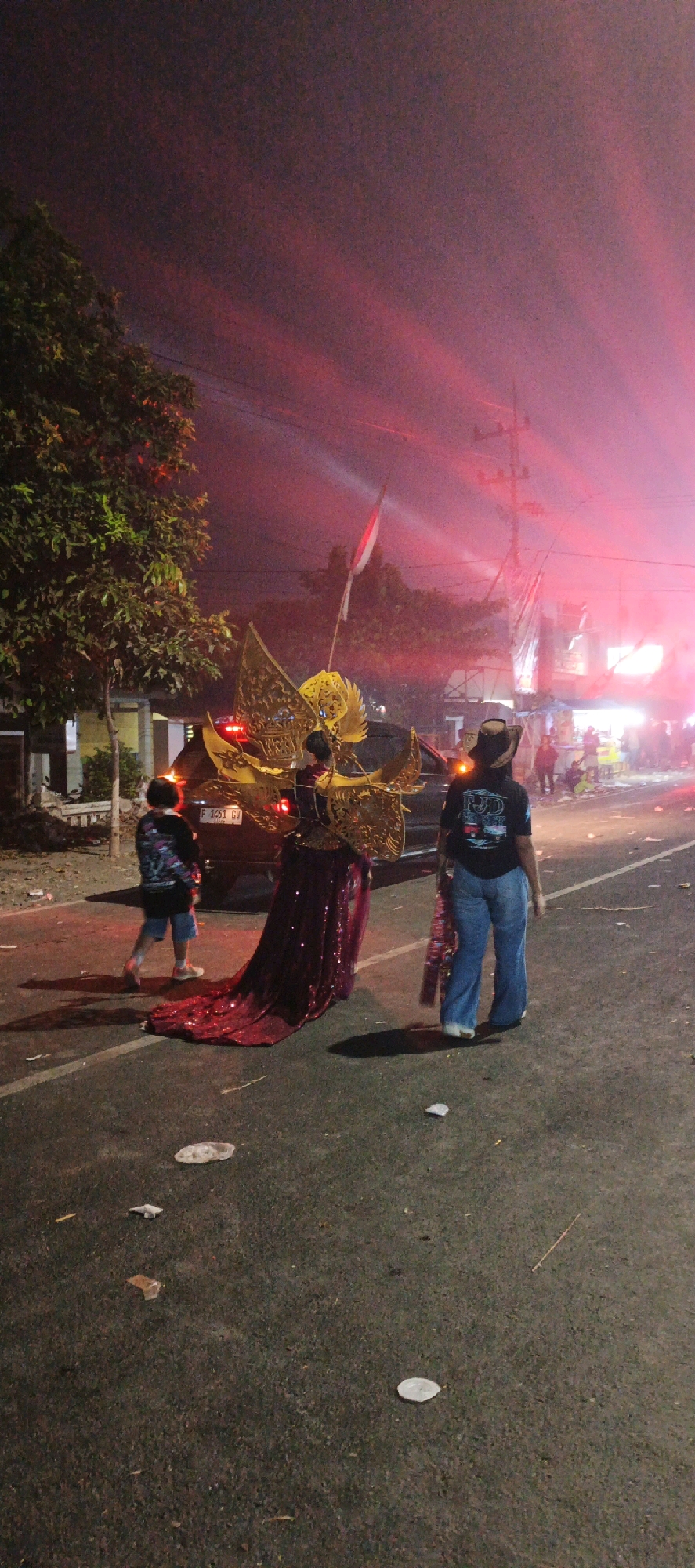 In Kasiyan, Indonesia, at night, a ceremonial parade is in progress on a paved street. A person in the foreground, viewed from behind, wears a black t-shirt and blue bell-bottom jeans, with a cowboy hat on their head. To the left, another person, also viewed from behind, is dressed in a black t-shirt and blue shorts. The central figure is adorned in a striking costume featuring large, golden wings and a long, sequined, deep red gown. They stand beside a black car. The sky above is illuminated by red smoke, possibly from fireworks or flares, creating a dramatic atmosphere. A red and white Indonesian flag is visible on a pole in the background, and streetlights and buildings are dimly lit along the side of the road. The street surface shows scattered debris.