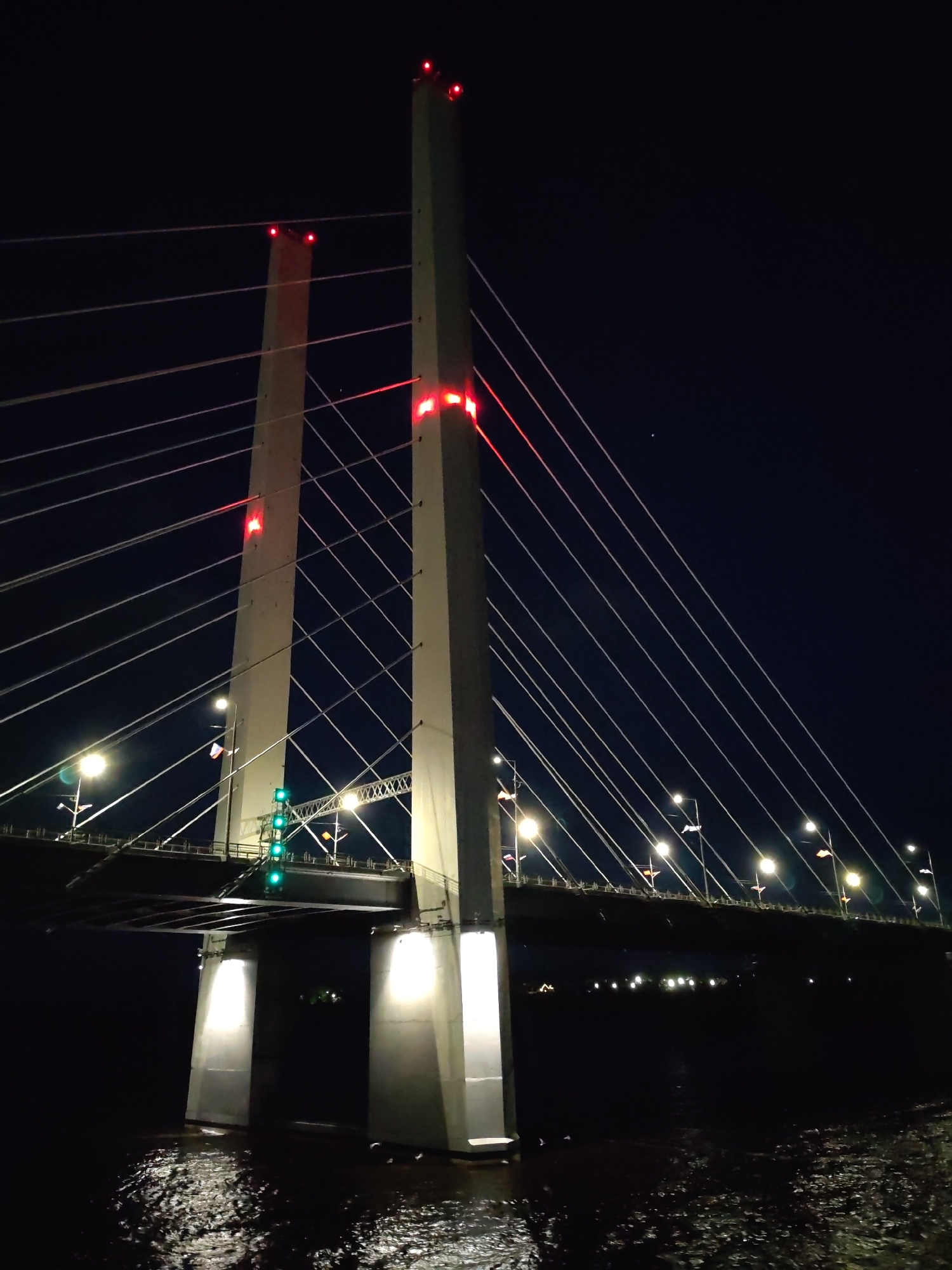 The image depicts a night view of the October Bridge in Cherepovets, Russia. The bridge is illuminated with bright white lights along its span and red lights atop the two prominent support towers. Multiple suspension cables extend from the towers to the bridge deck. The water reflects some of the light from the bridge. The sky is a dark blue. No people are visible in the scene.