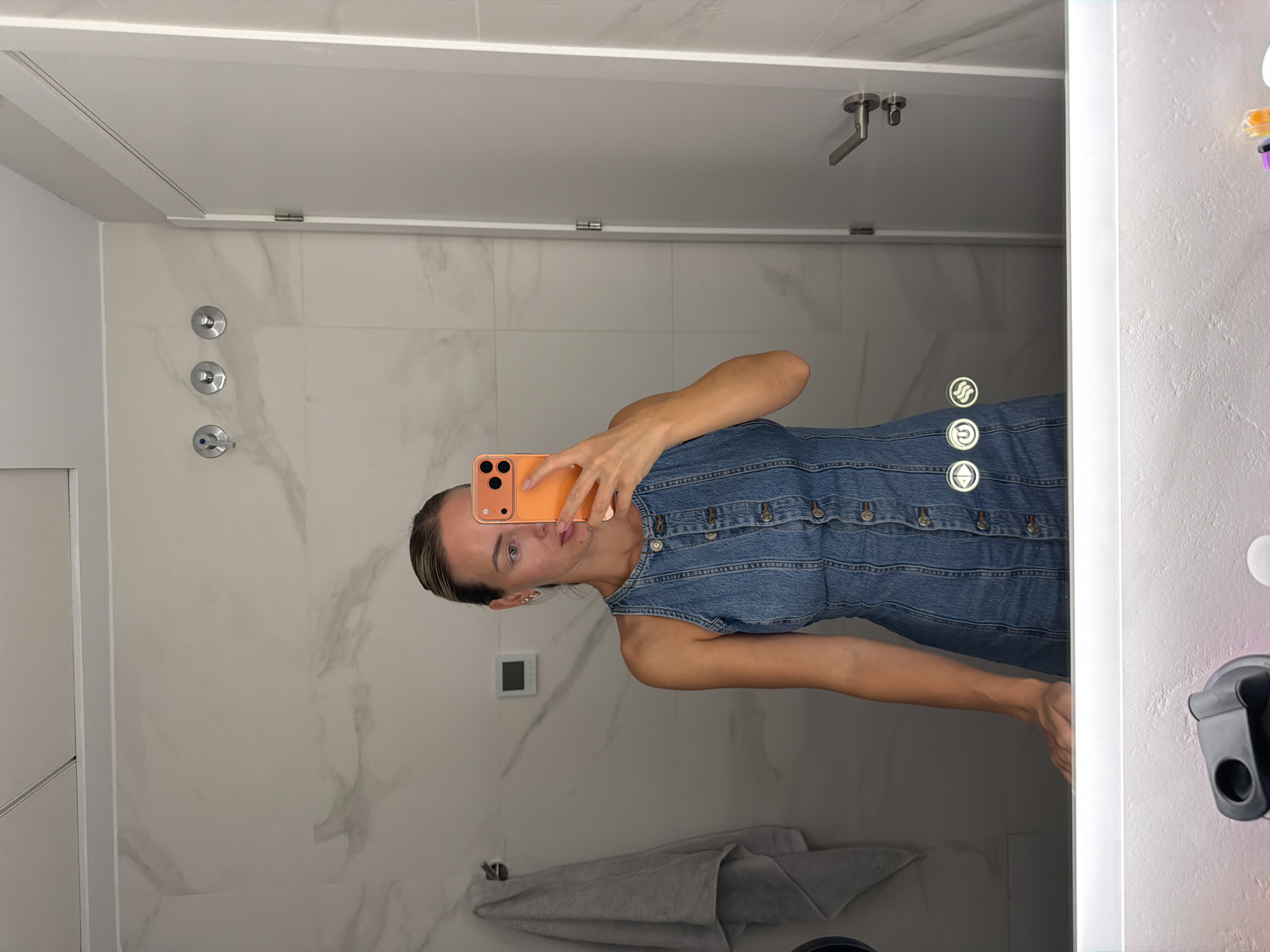 A blonde woman wearing a denim dress takes a mirror selfie in a modern bathroom. She holds an orange smartphone up to take the photo, which reflects in the mirror along with her face and upper body. The bathroom features white marble-tiled walls and a white door with a silver handle. A grey towel hangs on a rack to the left of the frame. The lower part of the mirror shows some icons illuminated, and the countertop below the mirror has a black object. The location is identified as Finestrat, Spain.