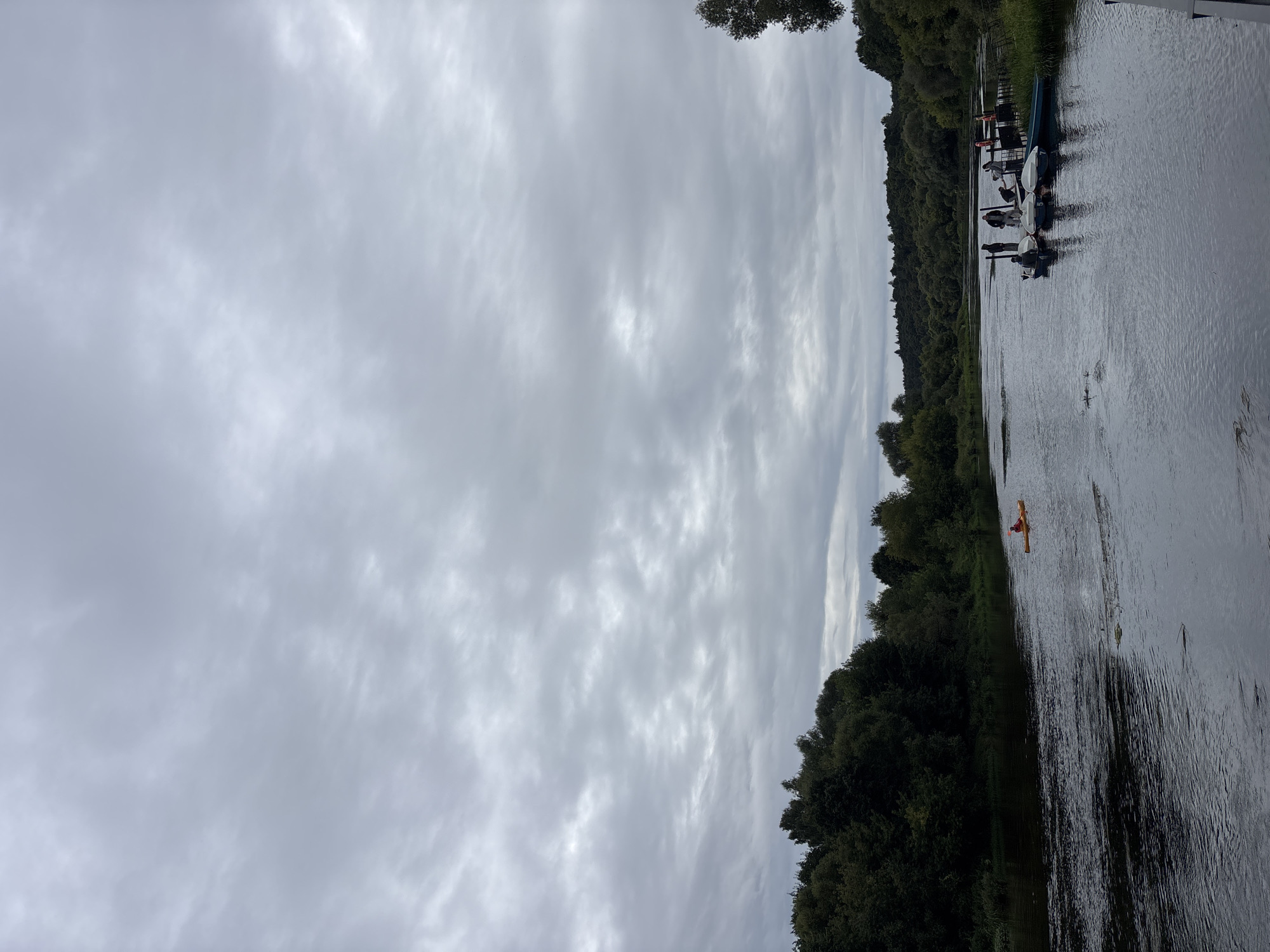 The image depicts a wide river in Zvenigorod, Russia, on an overcast day. The riverbanks are densely lined with green trees. In the foreground, the water is dark and rippled. On the left side of the river, a lone kayaker in an orange kayak is paddling. On the right side of the river, a dock is visible with several small boats moored to it. Several people are standing on the dock and near the boats, some appearing to be interacting with the boats or each other. The sky is heavily clouded with a muted gray tone.