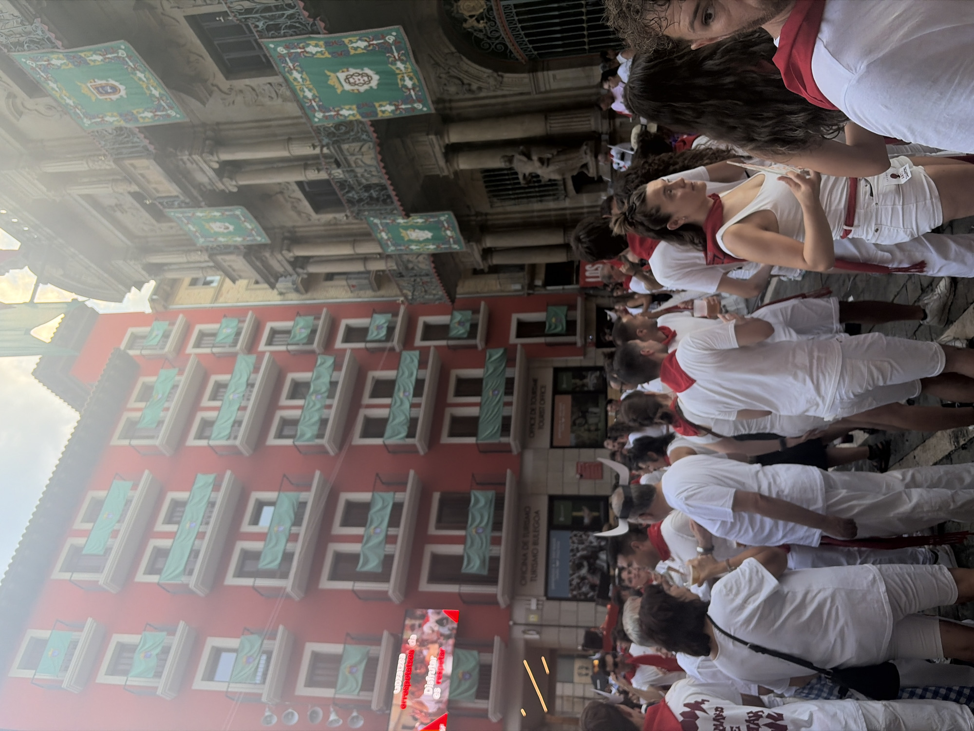 The image depicts a street scene in Pamplona, Spain, during the San Fermín festival. A large crowd of people, predominantly dressed in white with red sashes and neckerchiefs, are gathered in a narrow street lined with historic buildings. One building is painted a vibrant red with numerous balconies adorned with green banners featuring a coat of arms. Adjacent to it, a more ornate building with classical architectural elements also displays similar green banners.
In the foreground, several individuals are visible, some interacting with each other. One woman is holding a drink, and others are looking in different directions. Behind the crowd, a large digital screen is mounted on the red building, displaying what appears to be a live broadcast or recorded footage, possibly related to the festival events. The atmosphere seems festive and crowded, typical of a major public event in a European city. The presence of bull horns on a person's head further suggests the connection to the San Fermín bull-running festival. The overall lighting indicates it is daytime, with some atmospheric haze possibly due to the weather or smoke.