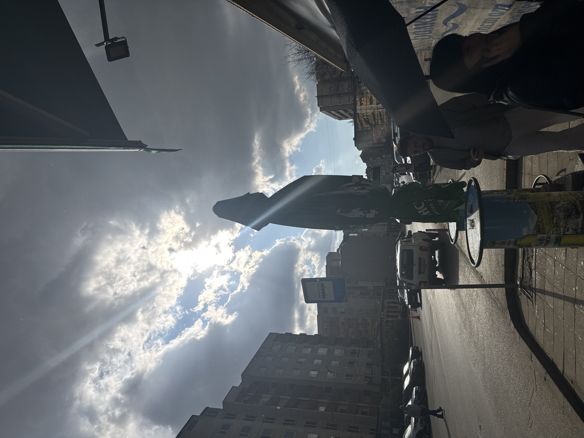 The image depicts a street scene in Sofia, Bulgaria. In the foreground, two people are seated at an outdoor table, partially obscured by an umbrella. A decorative trash receptacle stands nearby. The street is lined with parked cars and buildings. A bus stop sign is visible in the middle ground. The sky is filled with clouds, and sunlight streams through the gaps.