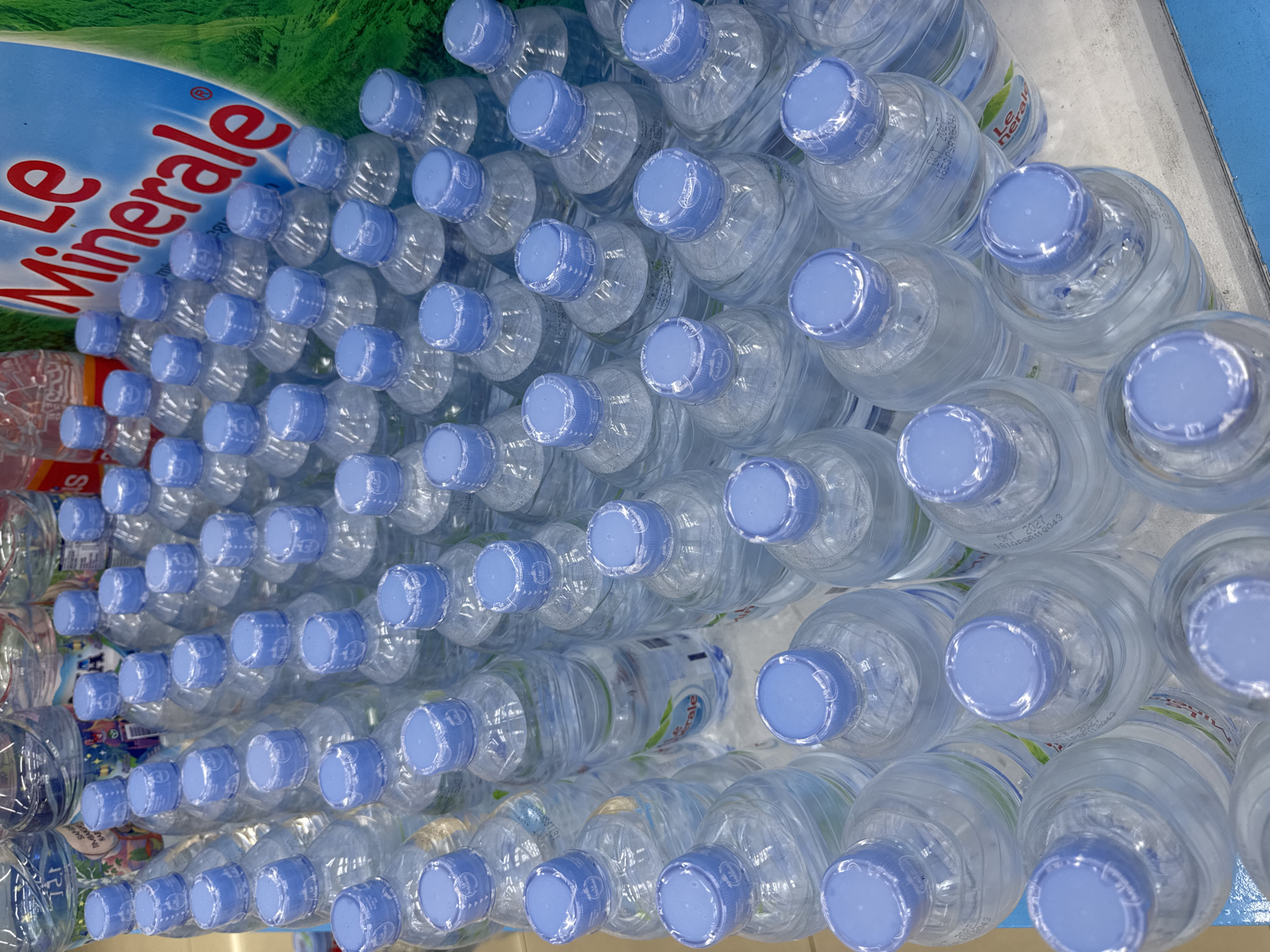 The image presents an elevated, slightly angled view of numerous bottles of "Le Minerale" brand water. The bottles are clear plastic with light blue caps and are densely packed together, suggesting they are likely stocked on a shelf or in a display. The bottles are arranged in rows and columns. The background includes the "Le Minerale" branding. The location is identified as Kuta Selatan, Indonesia.