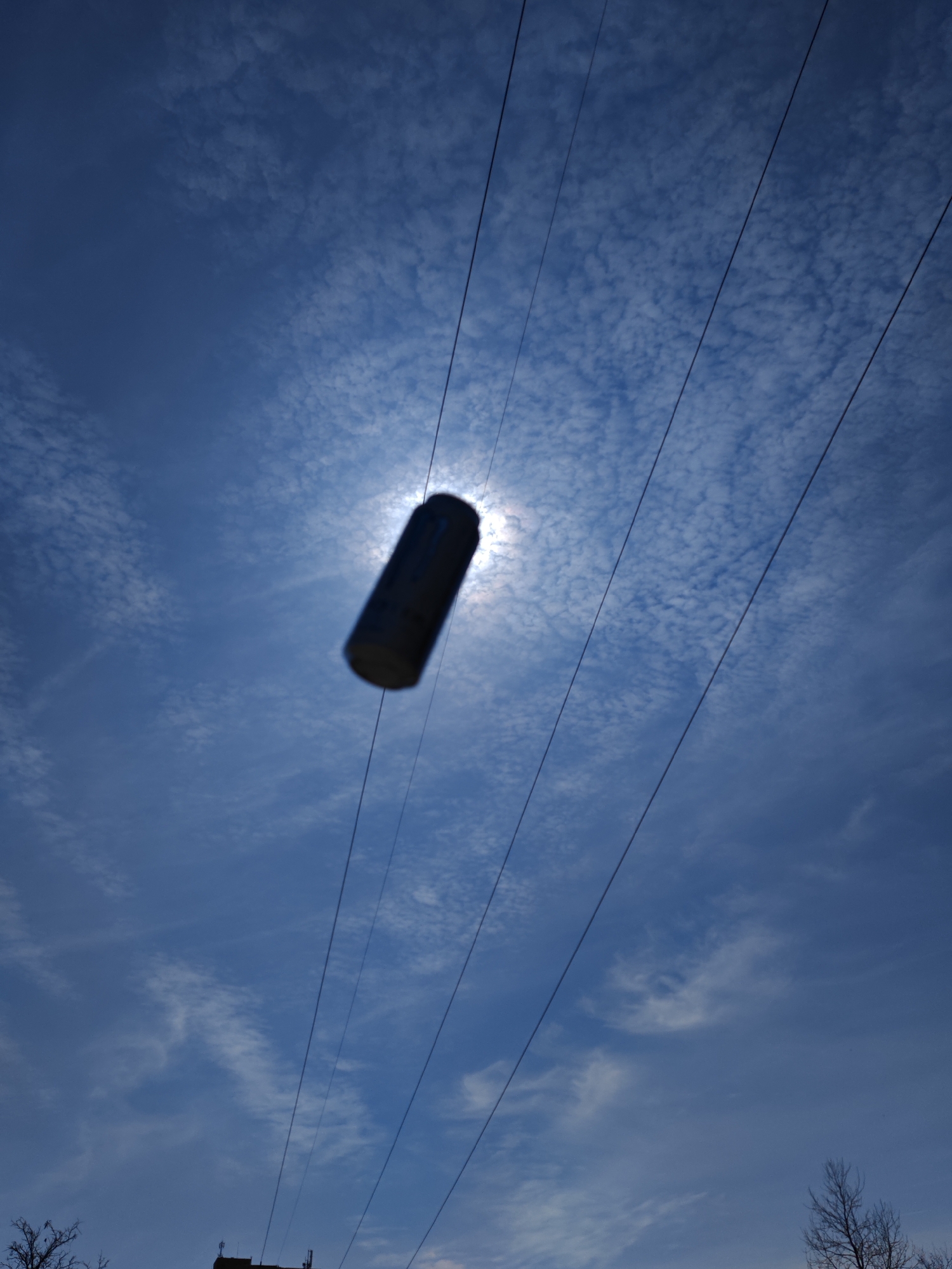 The image presents a daytime, upward-angled view of the sky over Shumen, Bulgaria. A deep blue sky with scattered white cumulus clouds fills the frame. The sun, partially obscured by a central dark, cylindrical object, emits a bright halo. The cylindrical object, consistent with a beverage can, is silhouetted and displays motion blur. Five dark, parallel wires or cables traverse the sky diagonally from the upper left to the lower right. In the bottom left, bare tree branches are visible. The silhouette of a building's upper section, possibly a rooftop with antennae, is discernible in the lower center-right.