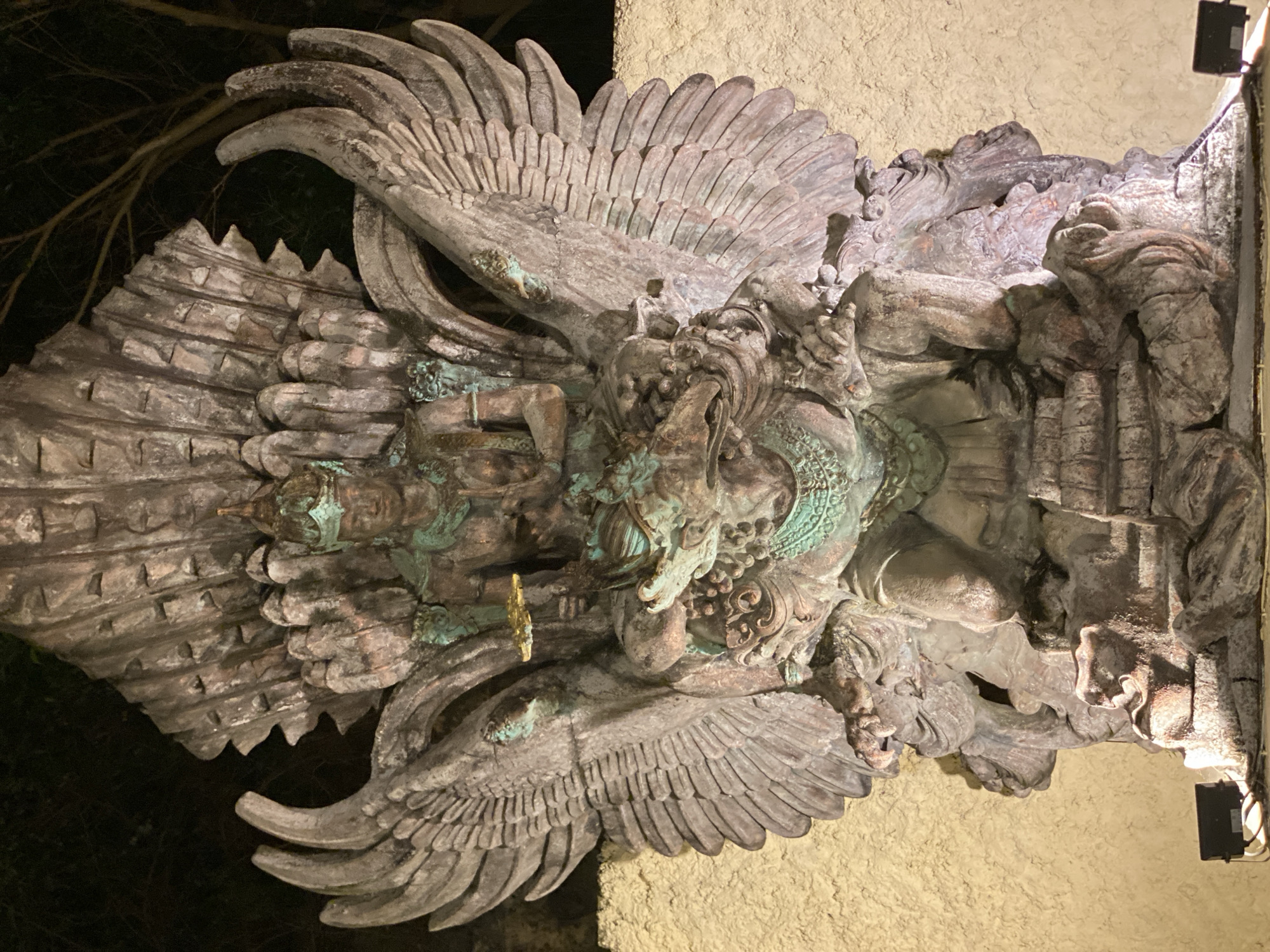 This is a vertical, close-up, full shot of a large, detailed stone sculpture depicting a winged mythical creature, possibly Garuda from Hindu mythology, adorned with multiple human-like figures integrated into its design. The sculpture is a weathered, dark gray stone, with significant areas of green patina, suggesting age and exposure to the elements. The creature's massive wings are spread wide, with each feather intricately carved. Within the body of the creature, there are several smaller, human-like figures, some with serene expressions and elaborate headdresses. One prominent figure appears to be in a seated or kneeling position. The sculpture is mounted on a textured, light-colored wall, which is illuminated by a warm light source, casting shadows and highlighting the texture of the stone. A dark, bare-branched tree is visible in the dark background on the left, indicating it's nighttime or a heavily shaded area. A small, black electrical box with wires is attached to the wall on the right, beneath the light source. The overall impression is one of grandeur and spiritual significance, common in Balinese art. The location context is Kuta Selatan, Indonesia.