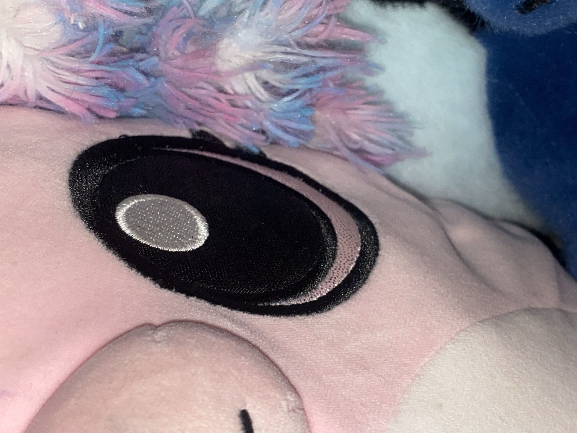 This is a close-up, eye-level shot of a pink plush toy, possibly a unicorn, with a large black eye. The eye is embroidered with a white oval shape inside, representing the pupil. The toy's fur is a mix of pink, blue, and white, with some strands appearing to have glitter. To the right of the eye, a white section of the toy is visible, which then transitions into a dark blue section. The overall impression is of a soft, cuddly toy. There is no discernible activity or event taking place, and no visible text. The lighting appears to be artificial, suggesting the photo was taken indoors. The focus is entirely on the toy's facial features and textured fur.