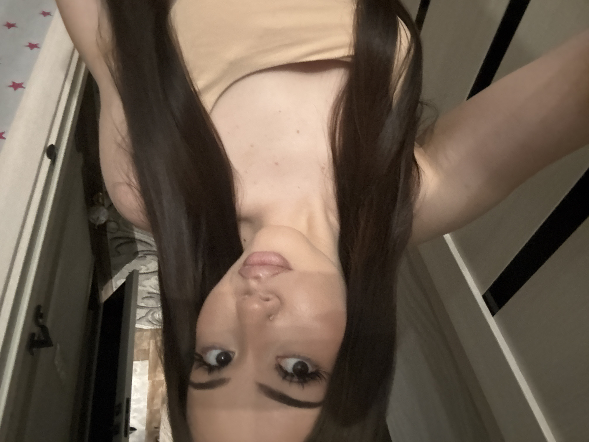 An inverted close-up selfie captures a person, likely female, with long, straight, dark brown hair framing her face. She is wearing a light-colored, possibly beige or tan, tank top or camisole. Her head is tilted slightly, and she looks directly into the camera with wide, expressive dark eyes, enhanced by prominent eyelashes and well-defined eyebrows. Her lips are full and slightly parted, showing a subtle pink tone. A small silver stud is visible in her nose piercing.
When viewed upright, the background reveals an indoor residential setting. To the left of the person's head, there's a light-colored surface adorned with small red star patterns, possibly a bed sheet, duvet cover, or wallpaper. A dark handle or knob is visible on what appears to be a door or piece of furniture. To the right, a wall or furniture item features distinct vertical dark stripes against a lighter background. Other elements in the blurry background suggest a personal room, possibly a bedroom, with glimpses of patterned wallpaper and a slightly ajar door or narrow opening. The lighting appears soft and ambient, typical of an indoor environment. The individual's expression conveys a sense of direct engagement, possibly playful or surprised. There is no visible text in the image.