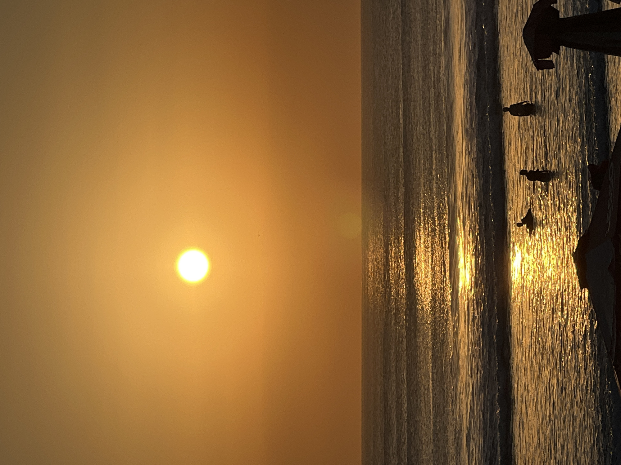 The image depicts a sunset at Kuta Beach, Bali, Indonesia, casting a warm orange glow over the scene. The sun is a bright, distinct orb in the upper center of the frame, its light reflecting off the water in a shimmering path. Several people are visible in the shallow water. In the foreground, to the right, the dark silhouette of a beach umbrella stands out. In the mid-ground, three figures are wading in the water. One figure is further out, appearing to be partially submerged. Another figure stands upright, and a third is nearer the shore, facing away from the camera. In the background, the calm ocean stretches to the horizon, with subtle waves breaking closer to the shore. The sky transitions from a bright orange near the sun to a deeper, warmer hue towards the top of the image.