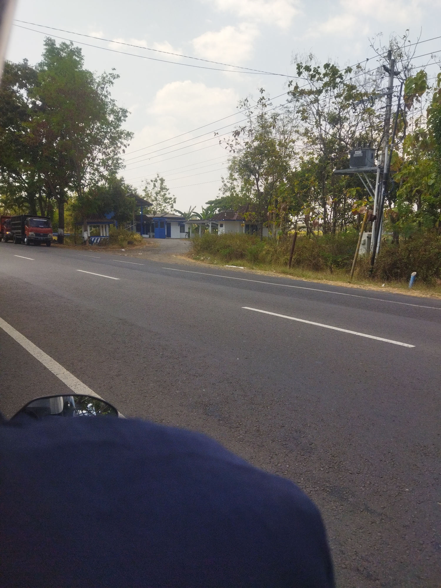 An eye-level, forward-facing outdoor shot captures a road scene in Nambangan, Indonesia. The asphalt road has a white dividing line. On the left side of the road, a red truck is parked. Further down the road on the left, a blue and white building with a small booth in front is visible. On the right side of the road, trees and vegetation line the roadside, with a utility pole supporting electrical equipment and wires. The sky above is overcast. The lower portion of the image shows a dark blue object, possibly clothing or a vehicle part, in the foreground, obscuring the bottom of the scene.