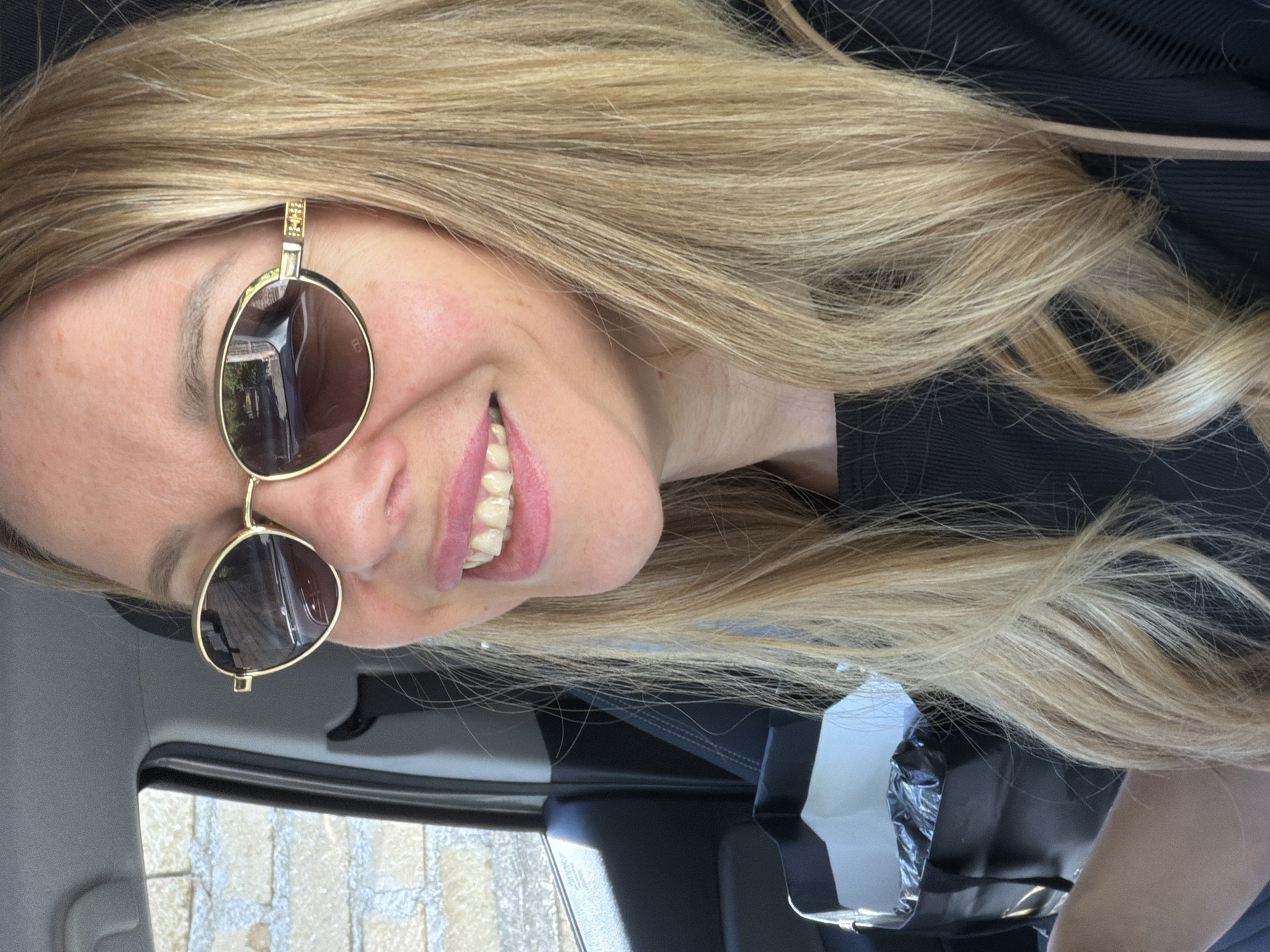 A close-up, waist-up, outdoor selfie shows a Caucasian woman with blonde hair and sunglasses smiling inside a vehicle. She is wearing a black shirt. A shopping bag is visible on her left. The background shows a brick wall and a car's interior. The location is not specified to be in Rome, Italy.