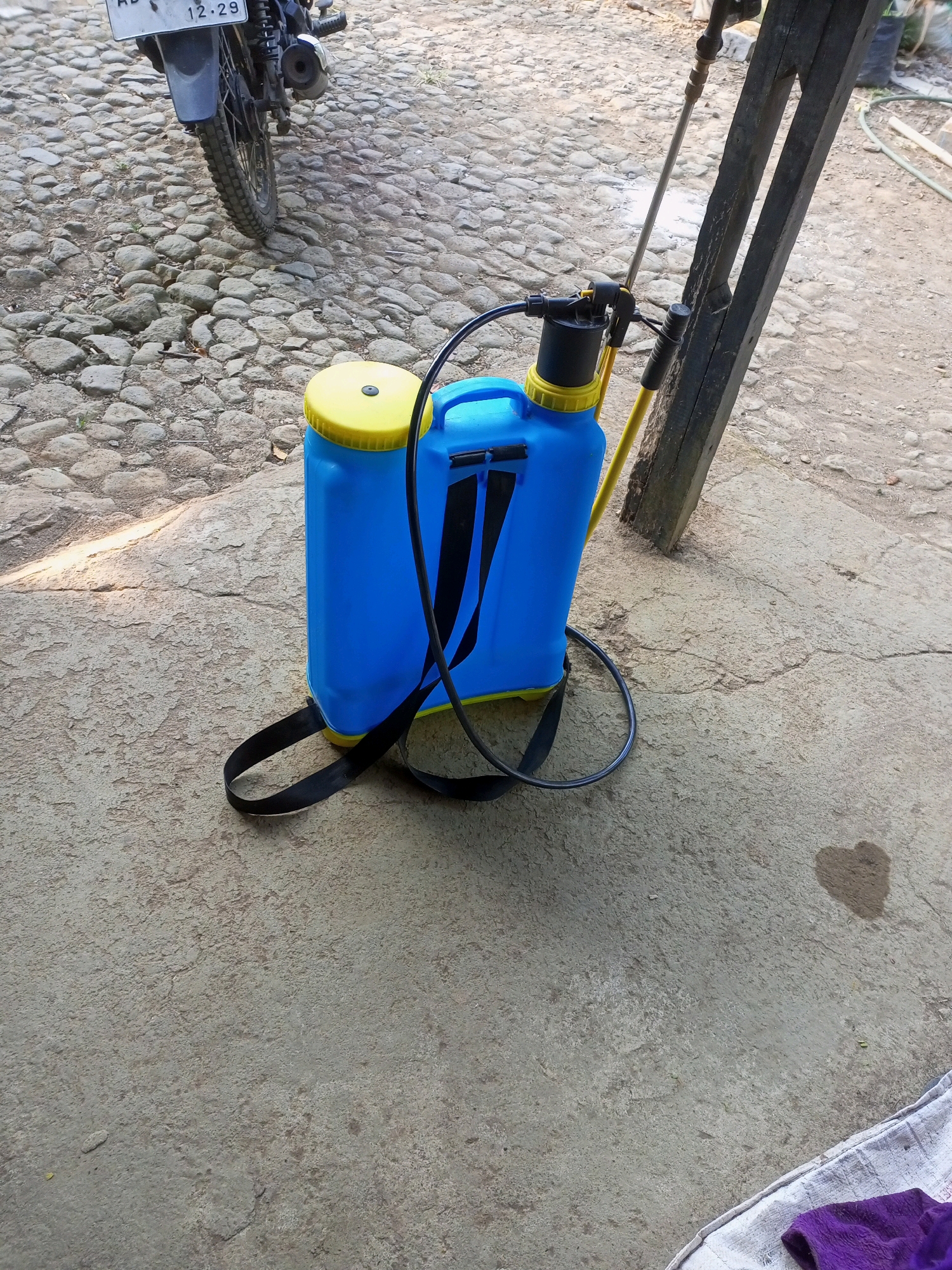 A blue backpack sprayer with a yellow lid and black straps is situated on a concrete surface. The sprayer is positioned next to a dark wooden beam. To the left of the sprayer, a portion of a motorcycle with a license plate reading "AD 12-29" is visible, resting on a cobblestone surface. The environment appears to be outdoors, possibly in a yard or street in Jatipurno, Indonesia, given the architectural style of the wooden beam and the cobblestone road. The concrete surface shows signs of wear and tear, with some textured areas and a heart-shaped water stain. A white cloth with purple fabric is present in the lower right corner of the frame.