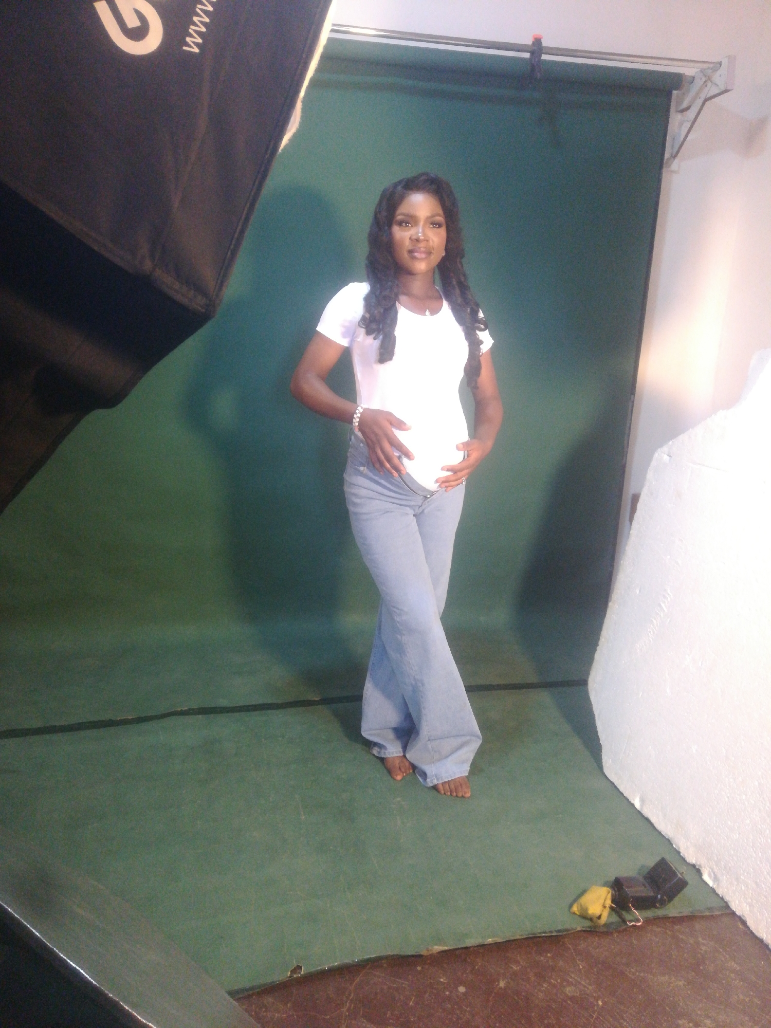 In an indoor studio setting with a green backdrop, a pregnant Black woman stands facing the camera. She is wearing a white t-shirt and light blue flared jeans. Her hands are resting on her pregnant belly, and she is wearing a bracelet on her left wrist. Her hair is long and dark, styled in waves. The floor of the studio is covered with a green mat. In the foreground, a large black photographic light modifier is visible on the left side, and a white styrofoam prop is on the right. Two small electronic devices, one yellow and one black, are on the floor near the bottom right corner. The overall lighting is bright, suggesting a professional photo shoot.