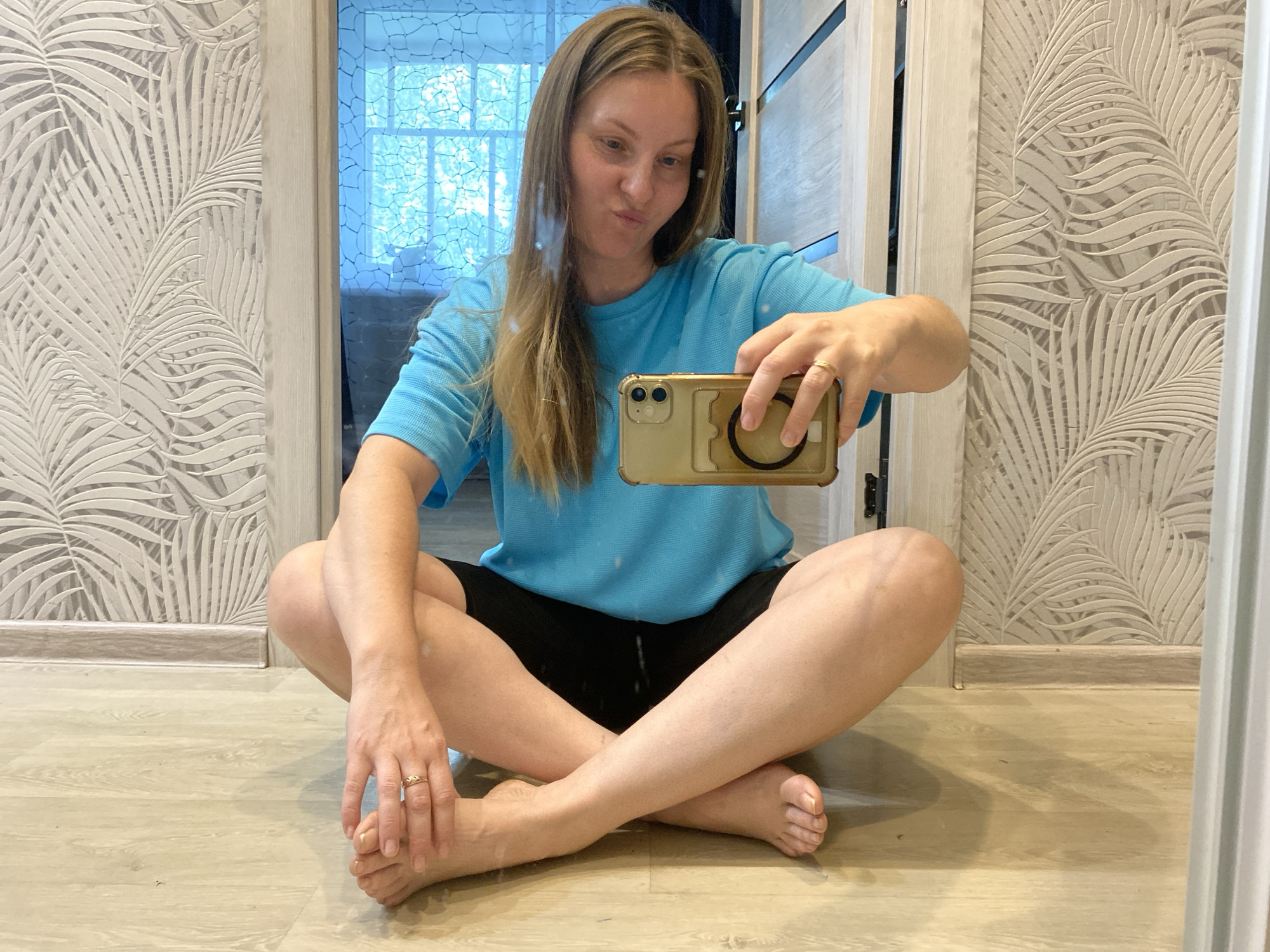 A fair-skinned woman with light brown hair is sitting cross-legged on a light-colored wooden floor, taking a selfie with a gold-colored smartphone. She is wearing a bright blue t-shirt and black shorts. On her left foot and hand, she is wearing a gold ring. Behind her is a doorway with a patterned wallpaper resembling palm leaves on either side. Through the doorway, a view of what appears to be a window with an abstract, mosaic-like pattern and a glimpse of the outside is visible. The photo appears to be taken in a room within a residence. There is no indication of the location being Noviy, Russia.