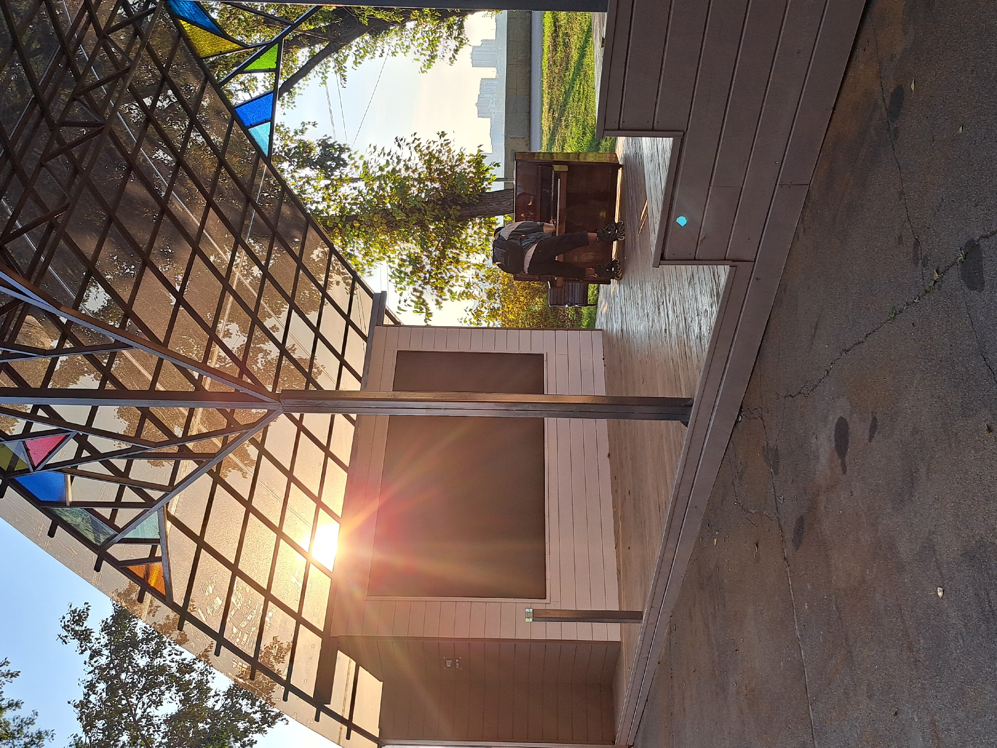 The image depicts an outdoor stage with a translucent, geometric roof structure adorned with stained-glass accents in blue, green, and orange. Sunlight streams through the roof, casting lens flares across the scene. A person wearing rollerblades and a backpack is standing in front of an upright piano on the wooden stage, appearing to interact with it. Behind the stage, trees with yellowing leaves are visible, along with a distant cityscape featuring apartment buildings. The ground in front of the stage is paved with cracked asphalt. The scene appears to be located in a public outdoor space, possibly a park or recreational area, in the city of Irkutsk, Russia.