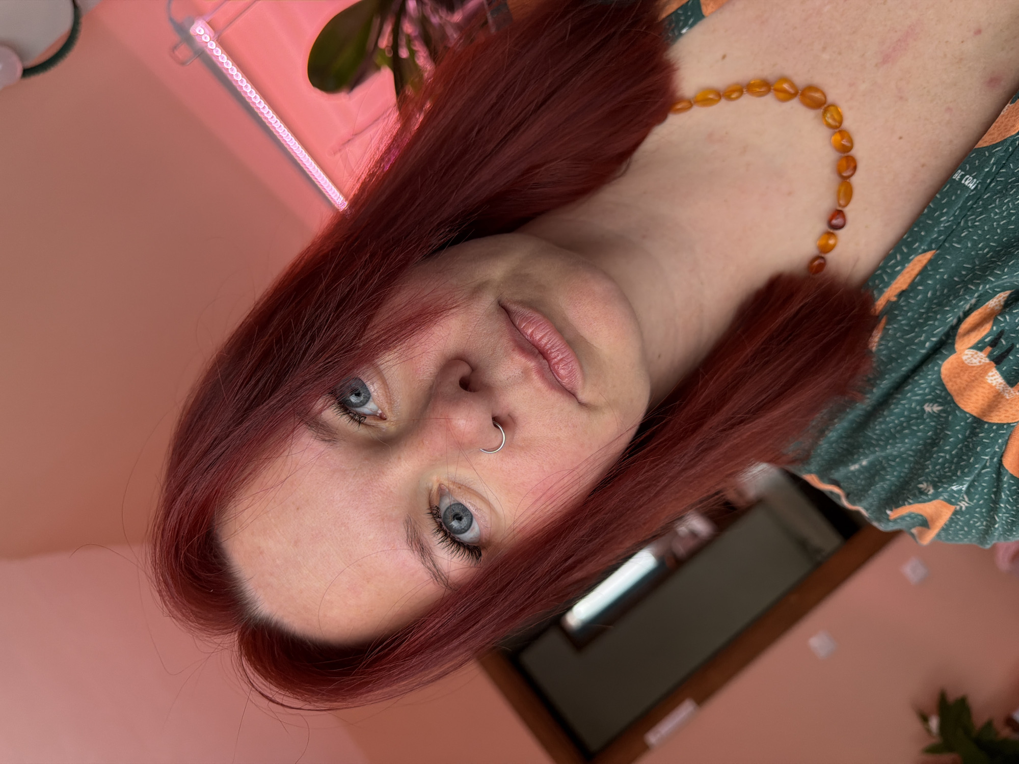 A tilted close-up shot of a woman with red hair and blue eyes. She is wearing a light green patterned top and an amber-colored beaded necklace. She has a silver nose ring. The background is a light pink wall, with a blurred glimpse of a window frame and indoor plants. The location is not discernible from the image.