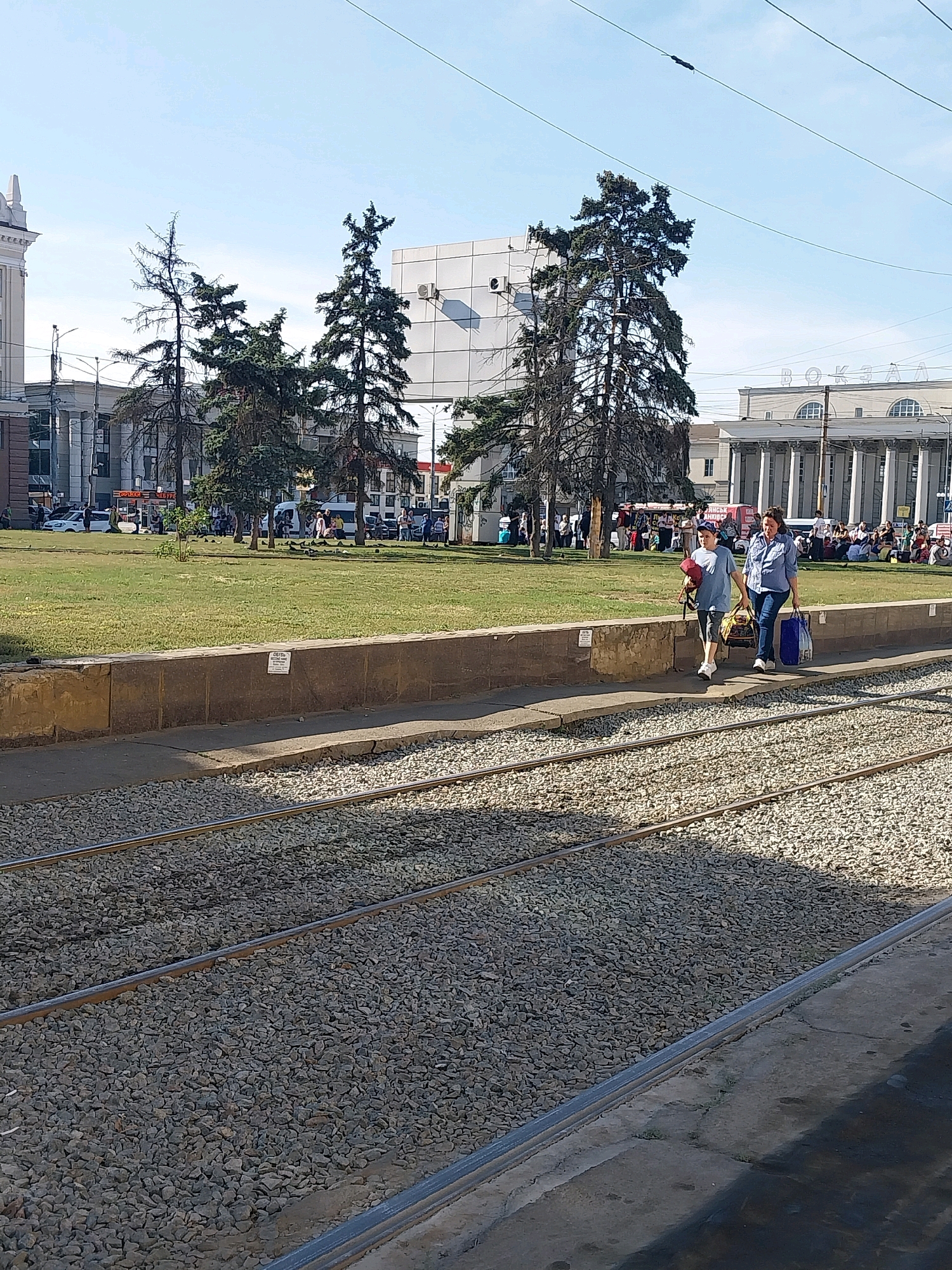 The image depicts a street scene in Dnipro, Ukraine, likely near a train station as suggested by the "BOK3AA" (вокзал - station) sign visible on the building in the background. In the foreground, train tracks with ballast are prominent. Two women are walking on a paved area beside the tracks, with one carrying a blue bag and the other a duffel bag and a red object. Behind them is a grassy area with several tall trees. Further in the background, there are buildings, including a large, classical-style structure with columns, and a modern building with a white facade. Several people are visible scattered in the background, some appearing to be gathered or waiting. Overhead, utility wires crisscross the sky. The scene appears to be under bright daylight.