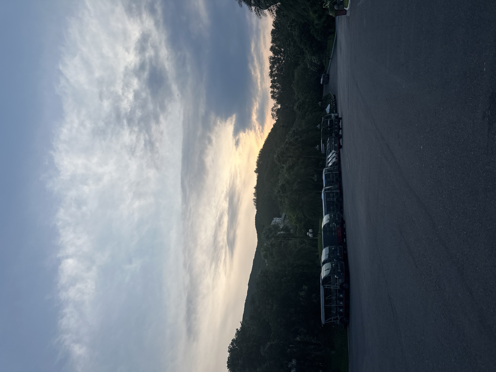 The image displays a serene landscape at either sunrise or sunset, with the sky dominating the upper portion of the frame. The sky exhibits a gradient from light blue to a mix of white and grey clouds, with a prominent warm, golden glow on the horizon, indicating the time of day. Below the sky, a range of hills or mountains is densely covered with lush, dark green foliage, appearing largely silhouetted against the bright horizon.
The foreground consists of a dark asphalt surface, likely a parking lot or road. Along the right side, a line of dark-colored vehicles is parked. These appear to be uniform, possibly mini-buses or shuttle carts, some with open sides. Text is visible on the side of several vehicles, clearly showing the word "Manoon".
The scene is calm and devoid of human activity. The soft, ambient light of dawn or dusk creates a peaceful atmosphere. The combination of mountainous terrain, dense greenery, and specific vehicle types suggests a location such as a resort, a large park, or a tourist area.