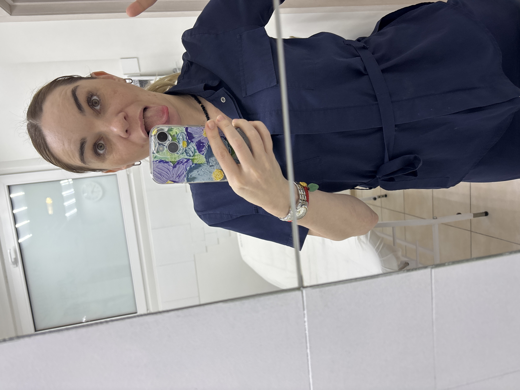 An image, rotated 90 degrees counter-clockwise, captures a woman taking a mirror selfie. She appears to be looking into her phone while holding it up to reflect her image. She has light hair, pulled back, and is wearing hoop earrings. Her eyes are open wide, and she has a playful expression, sticking her tongue out. She is dressed in a dark blue, long-sleeved shirt or dress, which features a tie-waist and possibly pockets on the chest. A black beaded necklace is visible around her neck, and on her left wrist, she wears a silver-toned watch with a red strap and a red bracelet with green and yellow accents. Her smartphone, an iPhone with two visible cameras, is encased in a colorful cover with a floral or abstract pattern in shades of purple, green, and yellow.
The background, visible in the mirror's reflection, depicts a sterile-looking room. White tiled walls are prominent, along with a large window or door frame with a white frame. Through the window's reflection, what appears to be lights or a clock can be seen. To the right, a white, minimalist bed or examination table with a metal frame is partially visible, indicating a potential clinic, spa, or professional treatment setting. The overall impression is casual and humorous, capturing a moment of lightheartedness. No legible text is visible in the image.