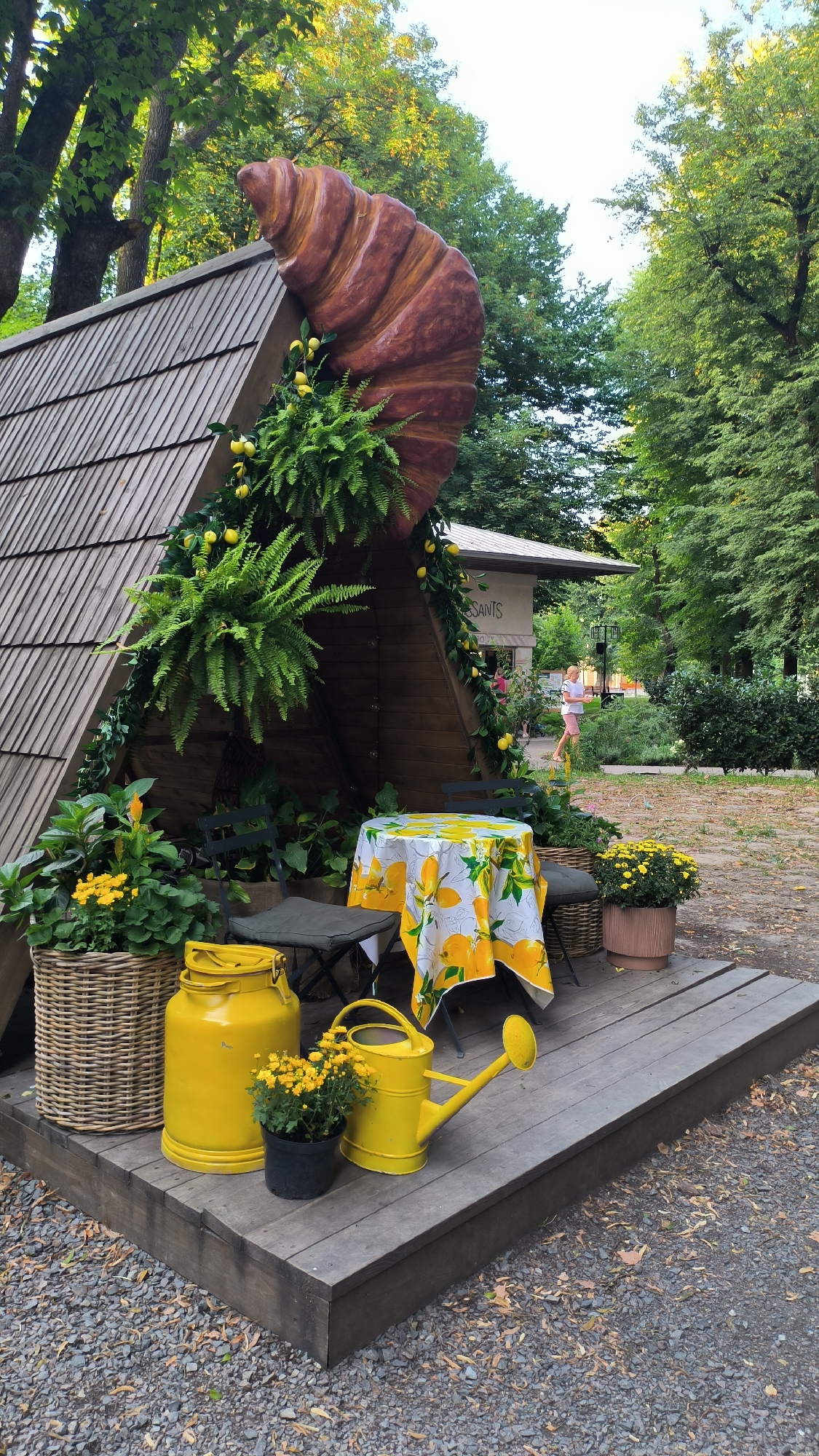 This image depicts an outdoor seating area decorated with a giant croissant sculpture on the roof of a wooden A-frame structure. The structure is adorned with greenery and faux lemons. A small table is set with a yellow tablecloth featuring a lemon print, two folding chairs, and is surrounded by potted plants and decorative yellow items including a milk can and a watering can. In the background, a building with a sign that appears to read "CROISSANTS" is visible, and a person in pink shorts and a white top is walking away from the camera. The setting appears to be a park or garden area, with trees and foliage surrounding the scene. The location is Lviv, Ukraine, likely a cafe or pastry shop.