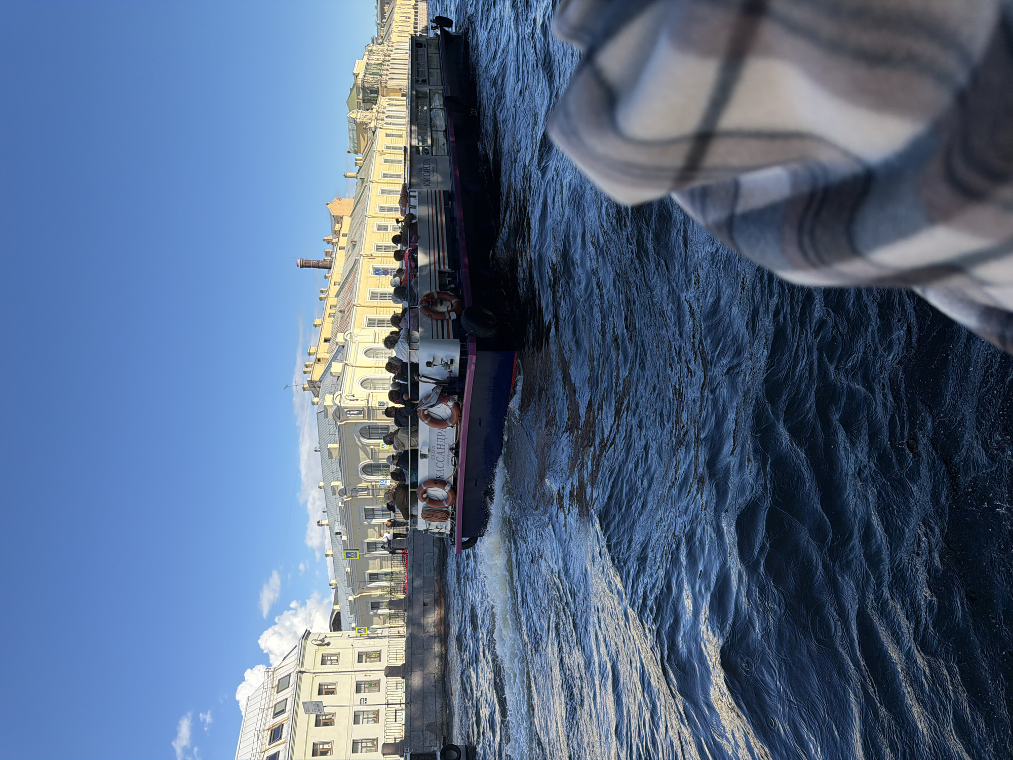 In a sunny, daytime scene in the city of Unknown, a boat cruise is underway on a canal. The boat, named "KASSANDRA," is filled with passengers seated on its open deck, many wearing casual clothing. Life preservers are visible on the side of the boat, suggesting a focus on safety. The boat is moving, creating ripples and waves in the dark blue water of the canal. In the background, a row of elegant, pale yellow buildings with white trim and numerous windows line the canal, reflecting the clear blue sky dotted with a few white clouds. A pedestrian bridge is also visible connecting the canal banks. The foreground is partially obscured by a close-up view of a checkered fabric, possibly a blanket or clothing, implying the photo is taken from another vessel or a close vantage point.