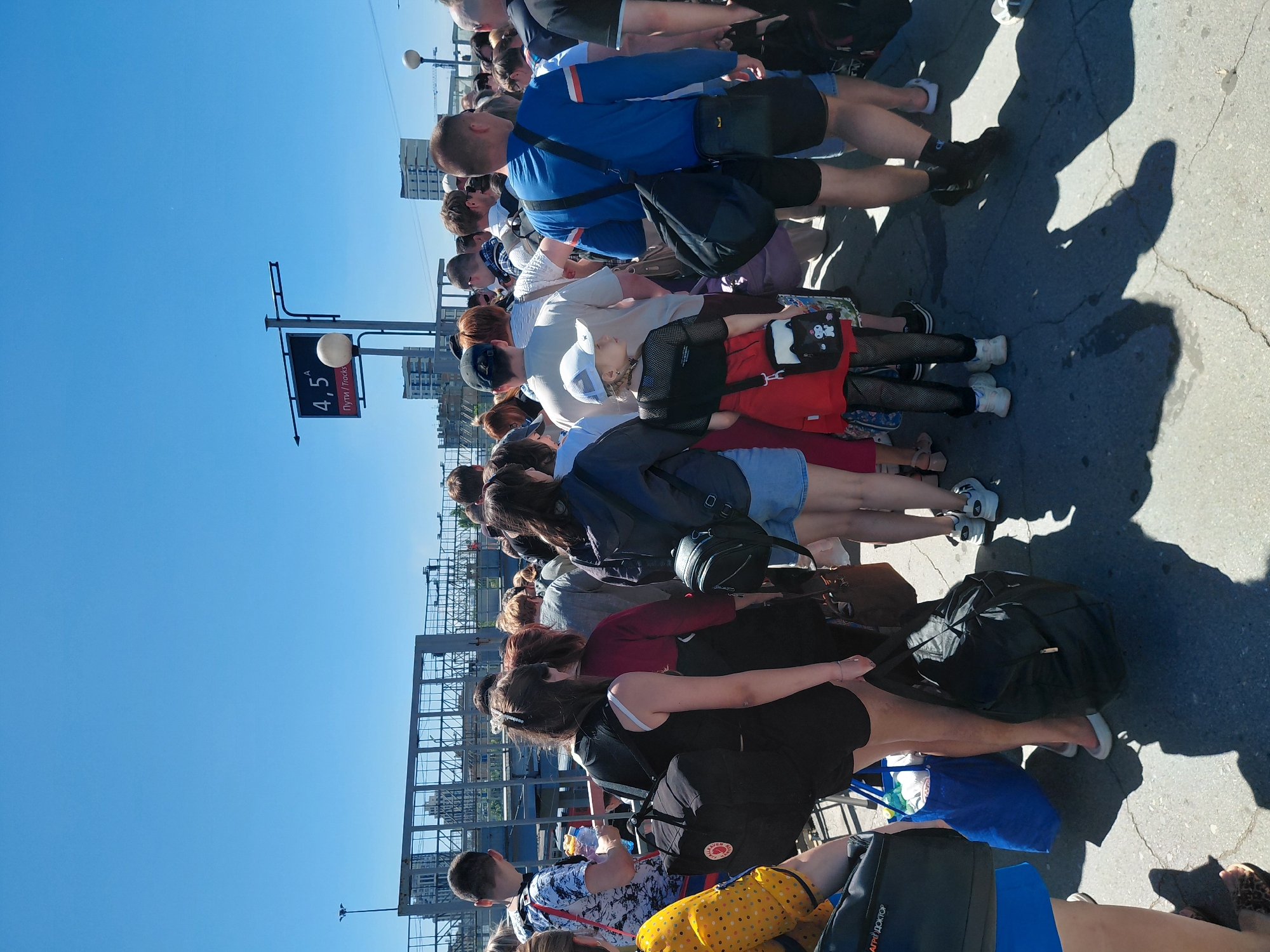 A group of people are standing in a line outdoors under a clear blue sky. A signpost indicates "4, 5A" and "Пути / Tracks" in Russian and English, suggesting a railway station. Many people are carrying bags and backpacks, with some wearing casual clothing like t-shirts, shorts, and skirts. The ground appears to be asphalt with visible cracks. In the background, there are buildings and industrial structures. The overall scene depicts a queue of passengers, likely waiting for a train.