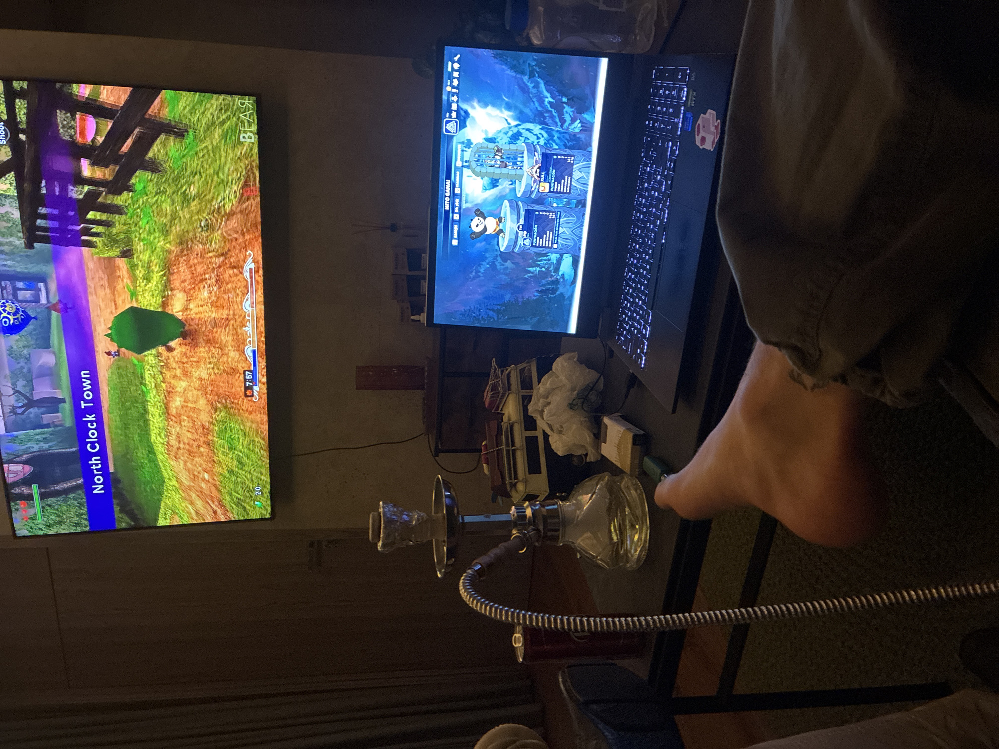 The image depicts an indoor scene at night, likely in a residential setting in Huixquilucan de Degollado, Mexico. A large flat-screen television is mounted on the wall, displaying a video game with the text "North Clock Town" visible. Below the television, a laptop computer is open on a table, also displaying a video game interface with characters and scores.
On the table, to the left of the laptop, is a hookah pipe with a glass base, metal stem, and foil-covered bowl. Next to the hookah is a red soda can. A packet of cigarettes and a lighter are also present on the table. A person's bare foot is visible in the foreground, resting on the edge of the table. The overall ambiance suggests a person is engaged in gaming and relaxation, possibly with a hookah. The room appears to be dimly lit, with the glow from the screens illuminating the immediate area.