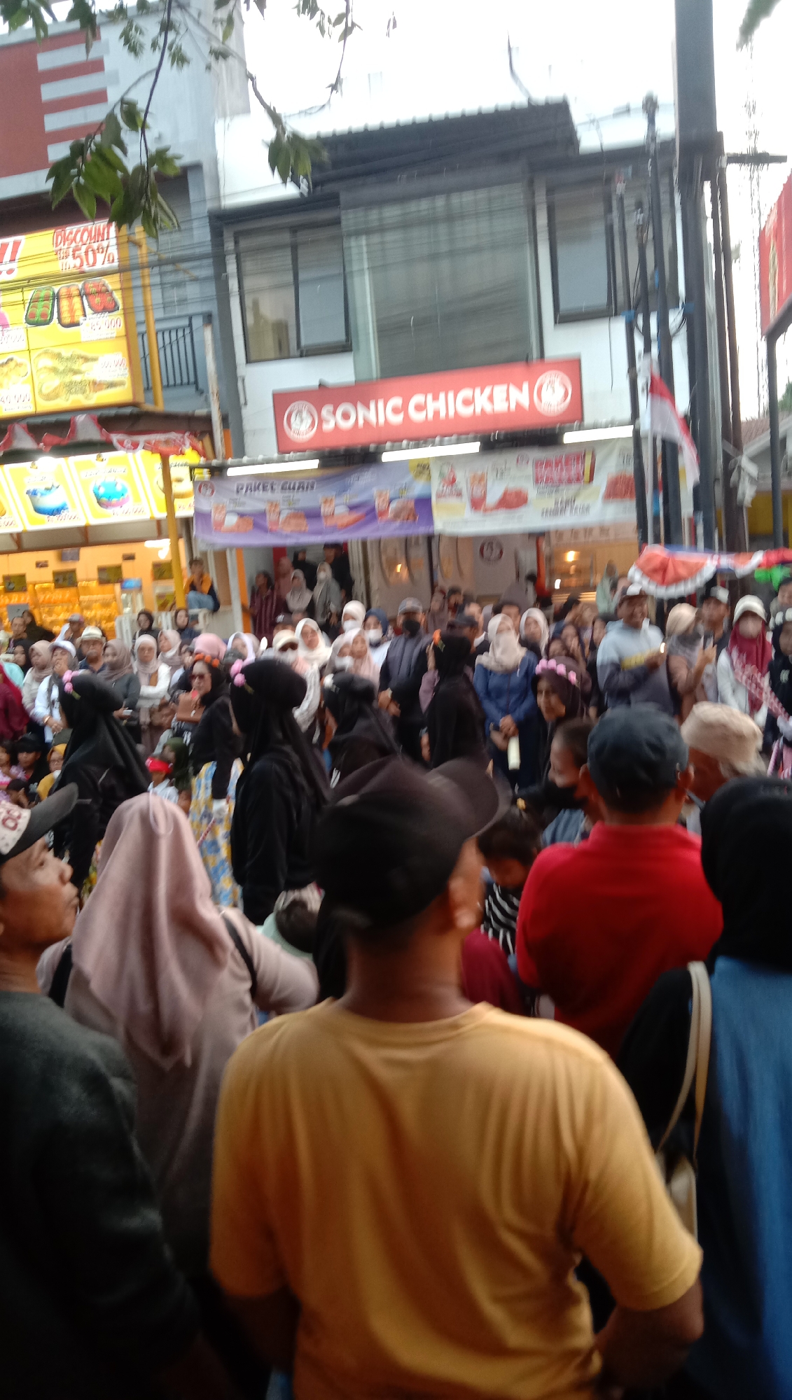 In the city of Jatisawit, Indonesia, a lively street scene unfolds in front of a Sonic Chicken restaurant. A large crowd of people, many wearing Islamic attire such as hijabs and veils, are gathered. The focus is on the restaurant's signage, a bright red banner with white lettering that reads "SONIC CHICKEN." Below this, another banner advertises "PAKET CUAN," suggesting a package deal. The environment appears to be an outdoor commercial area, with some trees and building facades visible in the background. The lighting suggests it could be late afternoon or early evening. The people are mostly facing towards the restaurant or looking in various directions, indicating a general gathering or perhaps a promotional event.