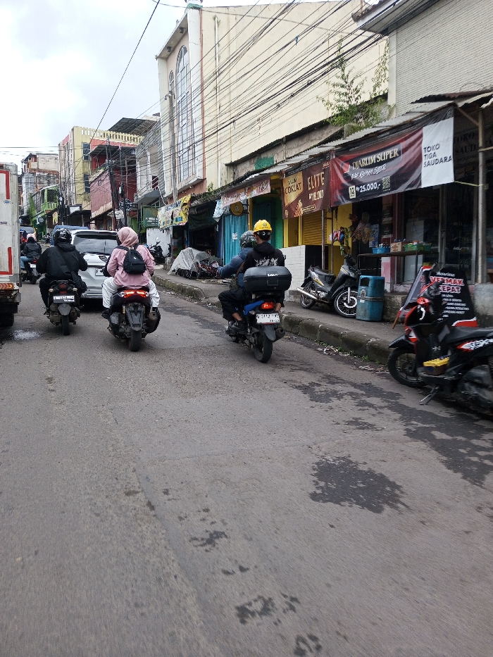The scene depicts an overcast daytime view of a street in a commercial district, specifically described as being in Melakudikadu, India. However, prominent signage displays Indonesian language, such as "DIARUM SUPER," "TOKO TIGA SAUDARA," "PROMO CIKOR," and "ELITE."
The asphalt road, appearing somewhat worn with dark stains and patches, shows two visible lanes of traffic moving away from the viewer. In the immediate foreground/midground, three motorcycles are in motion. From left to right:
1. A dark-clad individual wearing a dark helmet rides a dark motorcycle, partially obscured by a white truck on the far left.
2. A motorcycle with two occupants. The rider wears a pink head covering (hijab), a light-colored top, and white pants, carrying a black backpack. A passenger is seated behind this rider. The license plate appears to begin with 'B 30XX FPU'.
3. A single rider wearing a yellow helmet, dark top, and dark pants on a dark motorcycle, which has a black storage box mounted on its rear. Its license plate is visible as 'B 3713 PII'.
A white car, possibly an SUV, is visible behind the middle motorcycle.
On the right side of the street, a raised concrete curb separates the roadway from a narrow sidewalk lined with a series of single to two-story commercial buildings. These buildings feature various shopfronts, some with partially open or closed metal roll-down shutters. Several motorcycles and scooters are parked along the curb. One notable parked scooter is black with yellow accents. A blue cylindrical trash bin is visible on the sidewalk. Overhead, numerous electrical and communication cables crisscross the sky. In the background on the left, a taller, light-colored building with a distinctive arched window structure stands prominently among other multi-story buildings.