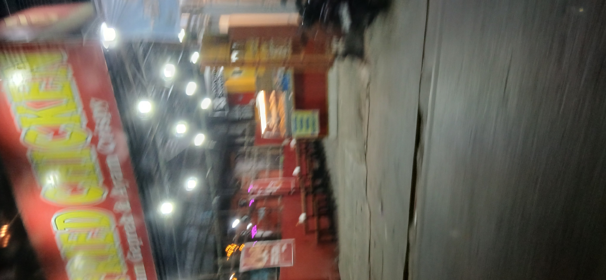 The image shows a blurry, nighttime scene of a fried chicken restaurant in Samarinda, Indonesia. A red banner with yellow lettering reads "FRIED CHICKEN" and below that, "Ayam Geprek & Ayam Crispy". The restaurant appears to have an open-air seating area with red tables and chairs. There are bright lights illuminating the establishment. In the background, a display case shows some food items. The street in front of the restaurant is paved and appears wet, suggesting it may have rained or is about to rain. A motorcycle is parked on the right side of the frame. The overall impression is of a bustling, local eatery in a Southeast Asian city.