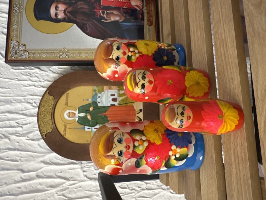 This image depicts an indoor scene in Zankovići, Montenegro, showcasing a collection of decorative objects positioned on a light-colored, slatted wooden surface. The background consists of a textured, off-white wall.
In the foreground, four traditional matryoshka dolls are arranged from left to right, decreasing in size. The largest doll, on the far left, has a blue base and a red body adorned with yellow, blue, and white floral motifs, featuring a painted face with black eyes and red lips. Immediately to its right is the second largest doll, similar in design with a blue base and red floral patterns. A third, smaller red doll with yellow floral patterns is situated to the front and right of the largest doll. The smallest doll, also red with yellow floral designs, is positioned in the leftmost foreground.
Behind the dolls, two religious icons are displayed. The icon on the left is a rounded rectangular piece with a brown frame, portraying a female saint in a white head covering and green cloak, holding a staff, with a white church building visible in the background against a golden sky. The icon on the right is larger and rectangular, featuring an ornate silver-toned border. It depicts a male saint with a long dark beard, wearing a dark monastic habit, with a golden halo around his head, and holding a red book. The scene is static, displaying a curated arrangement of these items.