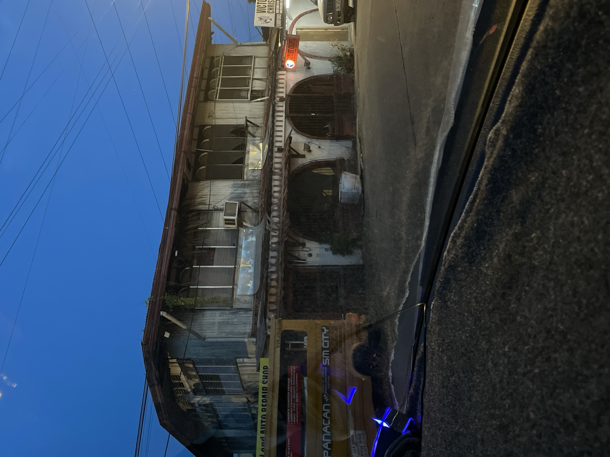 The image captures a street view in Davao City, Philippines, at dusk. The focal point is a weathered two-story building with a corrugated metal exterior. The ground floor features arched openings with metal grates, suggesting it might house shops or garages. A yellow van, identified with "PANACAN" and "SM CITY" branding, is parked in front of the building. The van appears to be an auto repair shop, as indicated by a yellow sign above it that reads "LAND AUTO REPAIR SHOP." Adjacent to the building, a sign for "VICTORY CHAPEL CHRISTIAN CENTER" is visible. The sky is a deep blue, transitioning from twilight. Overhead, a network of electrical wires crisscrosses the frame. The foreground shows the interior of a vehicle, with a dark, textured dashboard and a bright blue light emanating from a device on the left. The overall impression is of an urban commercial area in the Philippines, likely a neighborhood with a mix of businesses.
