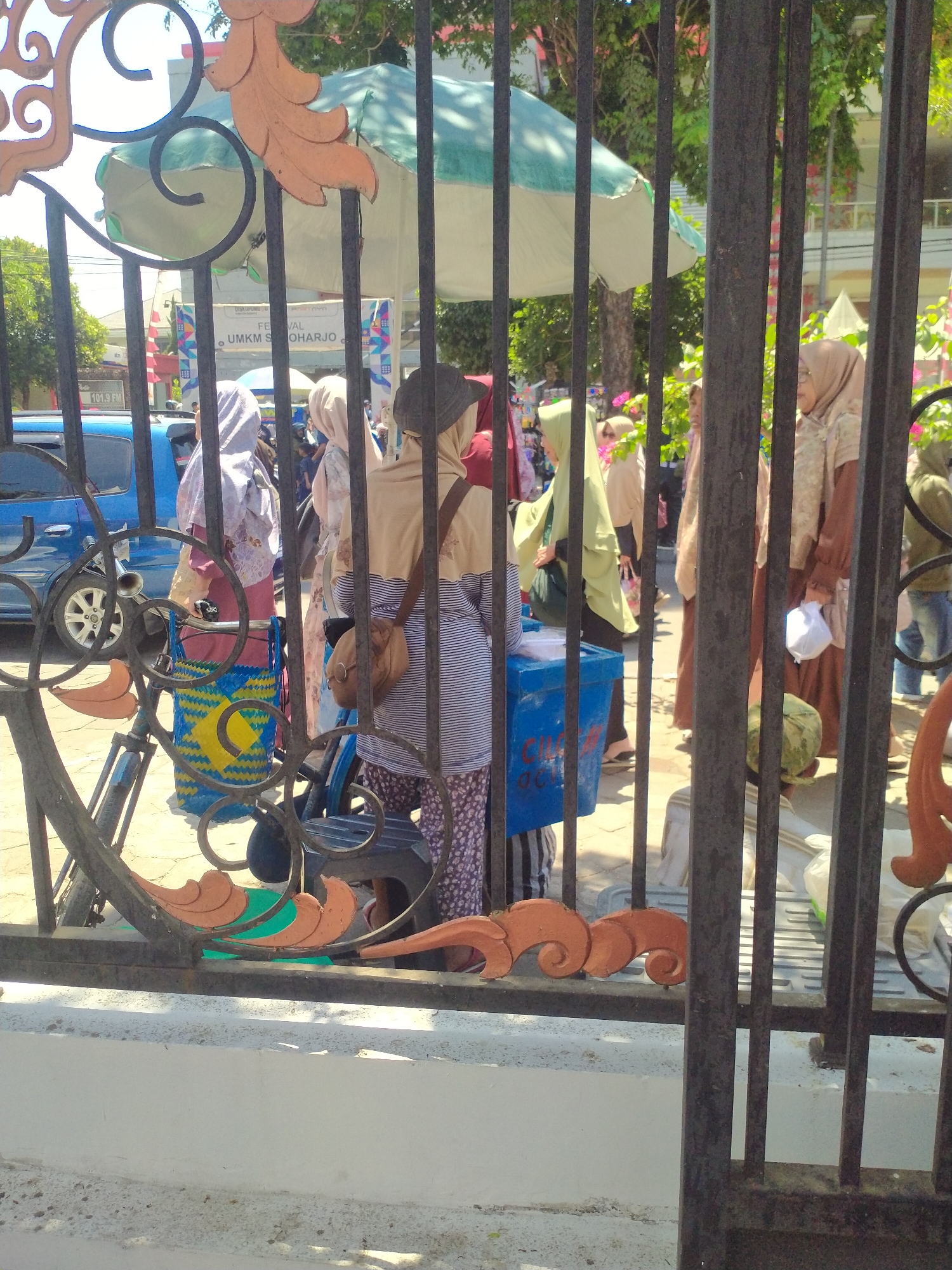 This image depicts a street scene in Sukoharjo, Indonesia, likely near a market or public gathering. The foreground is dominated by a black metal fence with ornate orange embellishments, partially obscuring the view. Behind the fence, several individuals, mostly women wearing hijabs, are visible. One woman in the center is wearing a striped long-sleeved shirt and patterned trousers, with a brown bag slung over her shoulder. To her left, another woman in a patterned dress carries a blue and yellow woven bag. In the background, a blue car is parked, and a green and white umbrella is set up. There's a sign visible that reads "FESTIVAL UMKM SUKOHARJO," indicating a local small and medium enterprise festival. The environment appears to be outdoors, with trees and buildings visible in the distance. The lighting suggests it is daytime, likely sunny.