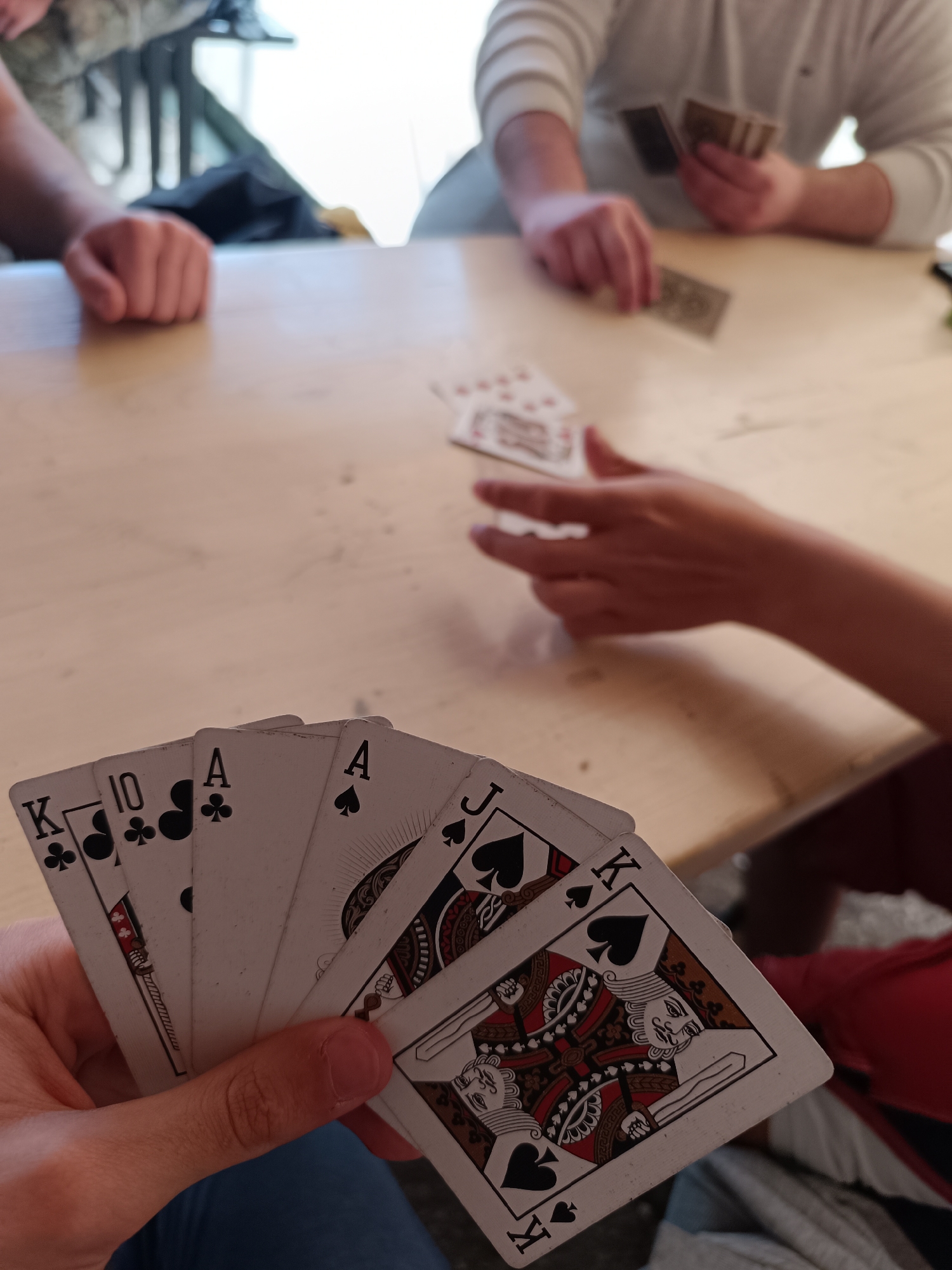 The image shows a group of people playing cards at a table. One person holds a hand of cards: King of Clubs, 10 of Clubs, Ace of Clubs, Ace of Spades, Jack of Spades, and King of Spades. Other players are present, with cards visible on the table. The location is possibly in Sofia, Bulgaria, but there is no definitive evidence of the specific city or country in the image itself.