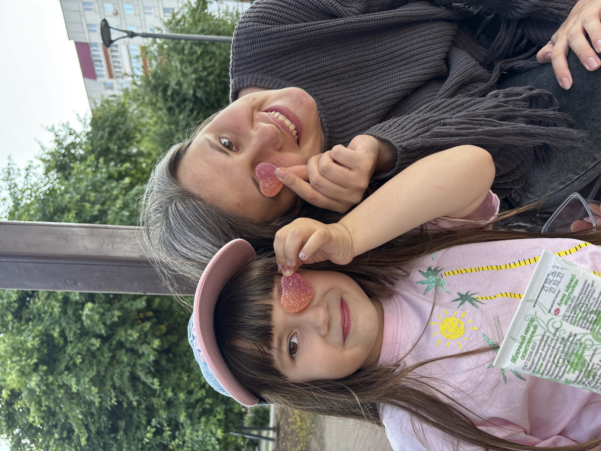 In Chaikovsky, Russia, two individuals, a child and an adult woman, are pictured outdoors. The child, wearing a pink t-shirt with a sun and palm tree design and a blue and pink baseball cap, holds a heart-shaped gummy candy over her right eye. The woman, with graying hair and wearing a dark gray sweater with fringe detail, also holds a similar candy over her left eye. Both are smiling and looking towards the camera. The background consists of trees and a multi-story building, suggesting an urban park or residential area.