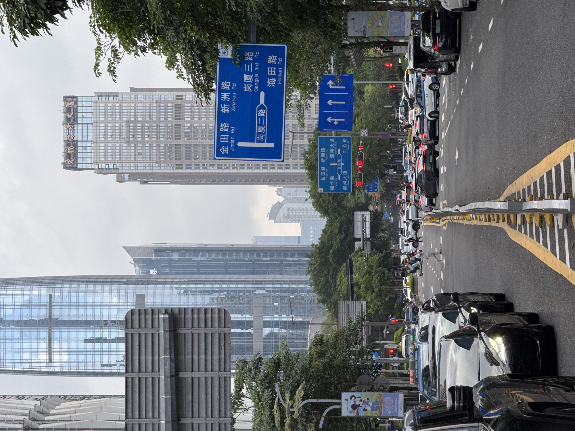 The image depicts a street in the Futian District of Shenzhen, China, filled with stationary traffic. Prominent buildings are visible in the background. Street signs direct traffic to Jintian Rd, Xinzhou Rd, Gangxia 3rd Rd, and Haitian Rd. There is also a sign for the Shenzhen Duty Free Business Building. A pedestrian is crossing the road. The sky is overcast.