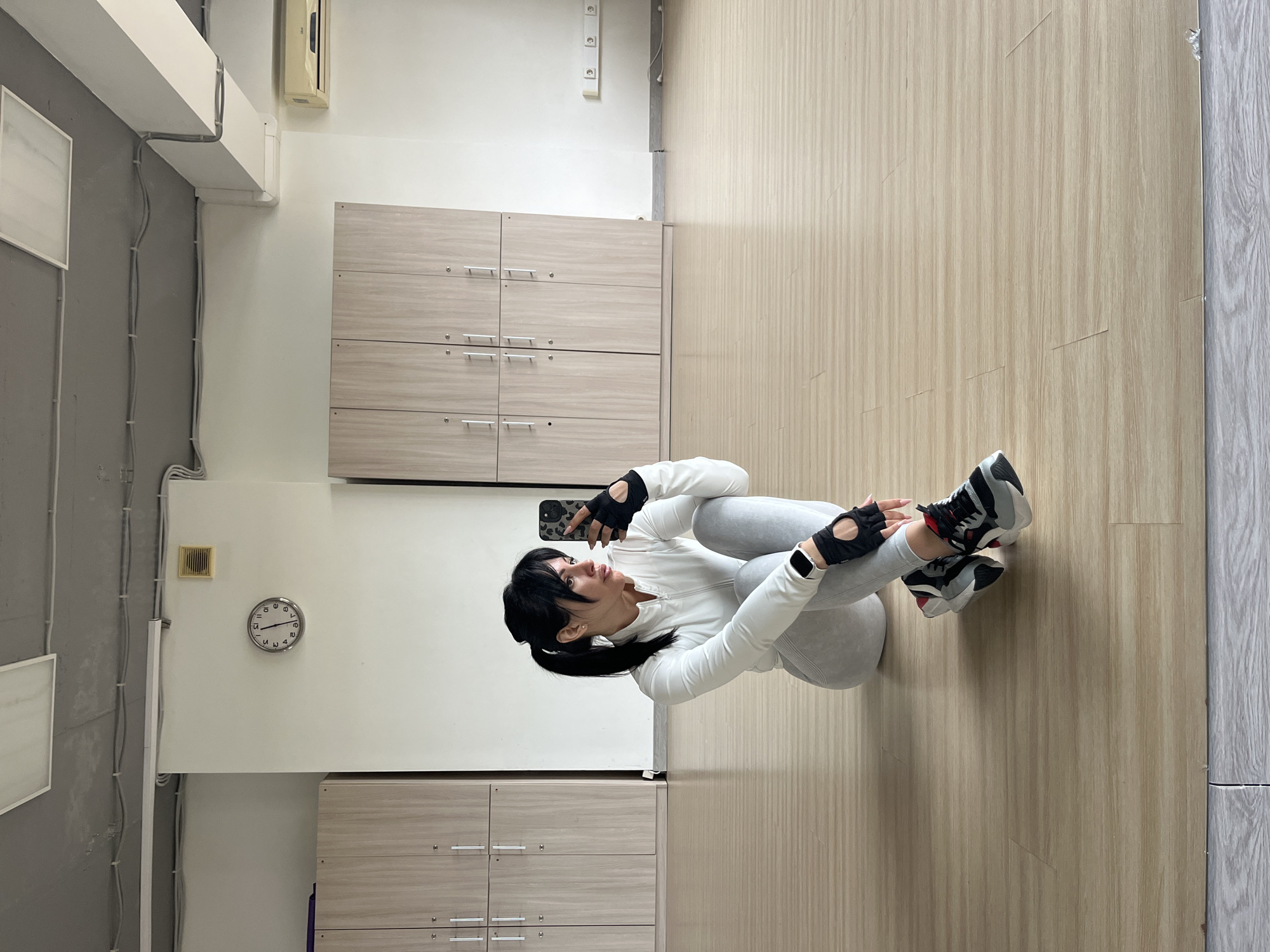 A woman is sitting on a gray fitness ball in a gym. She is wearing gray leggings, a white long-sleeved shirt, black gloves with finger cutouts, and gray, black, and red sneakers. She is holding a cell phone in her left hand and taking a selfie.
The gym has light wood-paneled floors and light wood-colored cabinets along the wall. A clock hangs on the white wall, and gray ventilation ducts and electrical conduits run along the ceiling. The location cannot be determined from the image.