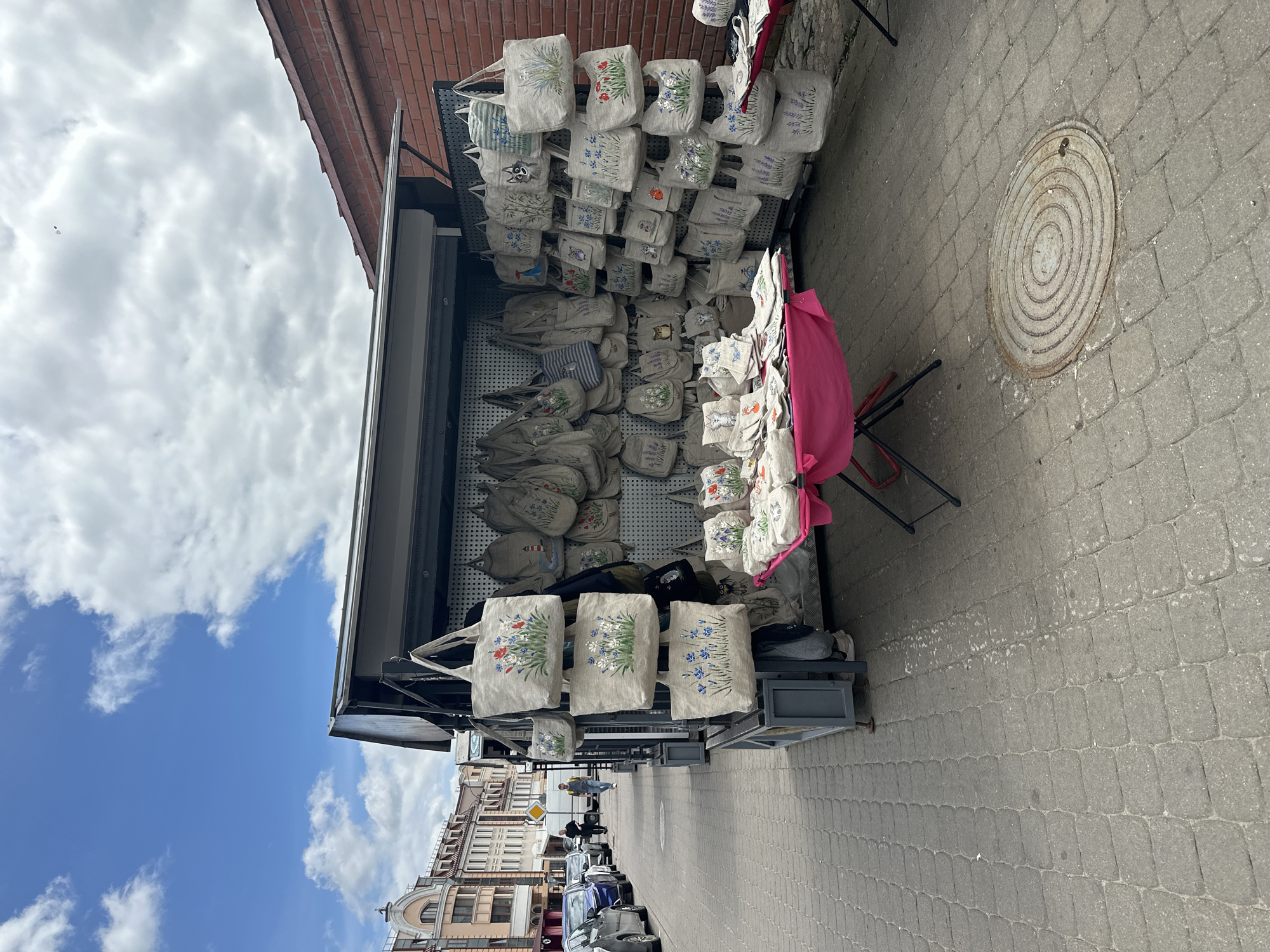 In Minsk, Belarus, a street vendor's stall displays an assortment of fabric bags, primarily made of linen, adorned with floral and animal motifs. The stall is set up on a cobblestone sidewalk adjacent to a street lined with cars and buildings. Several people are visible walking in the background, and the sky is partly cloudy. The bags are hung on a pegboard, with some displayed on tables covered by bright pink tablecloths. The overall scene depicts a typical outdoor market environment in a European city.