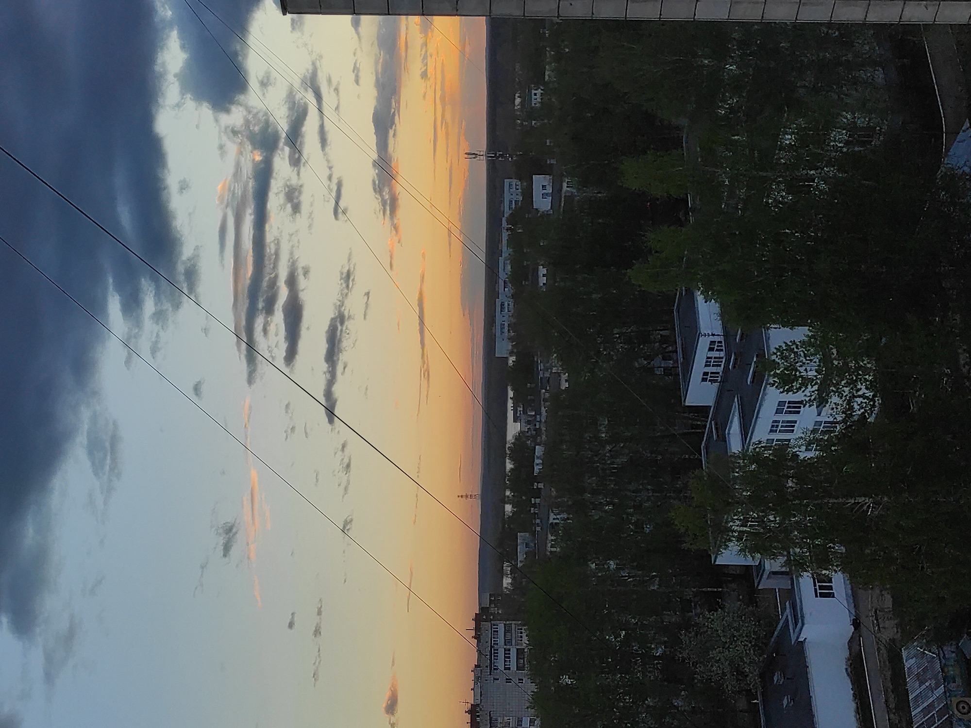 The image depicts a cityscape at dusk, likely in Chaikovsky, Russia, as implied in the prompt. The sky is divided between dark, stormy clouds and a warm, orange-tinged sunset. Power lines stretch across the sky. Below the horizon line, buildings and trees are visible. The buildings appear to be residential, with a mix of older and newer constructions. The trees are dense and predominantly green. The image is shot from an elevated perspective, possibly a high-rise building.