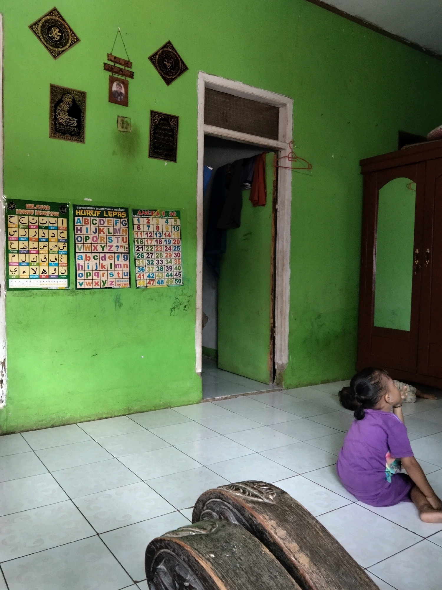 The image depicts an indoor scene, likely a home in Rahong, Indonesia. The walls are painted a vibrant green. On the left wall, there are several framed Islamic calligraphy and a small framed photograph. Below these, three educational charts are affixed to the wall: one with Arabic letters, another with English letters, and a third with numbers from 1 to 50. A doorway is visible in the center of the room, leading to another space. Clothes are hanging on a rack inside the doorway. To the right, a dark brown wooden wardrobe with a mirrored door stands against the wall. In the foreground, a young girl with dark hair tied in a bun is sitting on the tiled floor, wearing a purple shirt. She is facing away from the camera, with her head turned to the side. A wooden bench or table with carved details is in the lower left corner of the frame. The floor is covered with light-colored square tiles. The overall impression is of a simple, lived-in domestic space.