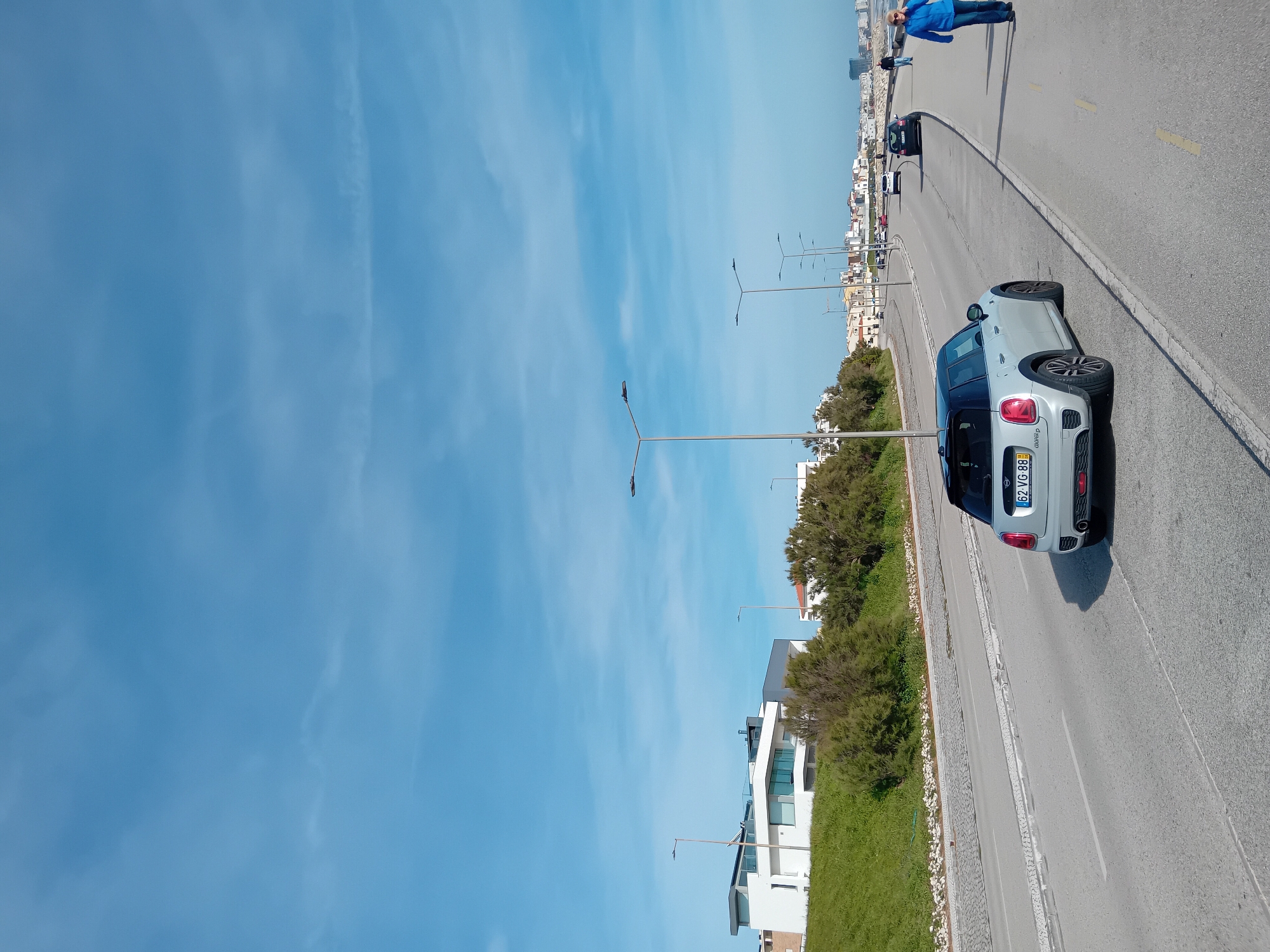 The image displays a sunny outdoor scene in Figueira da Foz, Portugal. The perspective is from the side of a paved road, which curves slightly to the right and recedes into the background. A light gray Mini Cooper car, with the license plate "62 VG 88" and "Coopera S" badging visible on the rear, is parked on the right side of the road, facing away from the viewer.
The road features a solid yellow line on its edge and a dashed white line, typical of road markings. Further down the road, a person wearing a blue jacket and dark trousers is seen walking, with another individual partially visible behind them. Other cars are present in the distance. Along the left side of the road, there are several modern, white multi-story buildings with large windows, interspersed with green grassy areas and dense green bushes. Streetlights are spaced along the roadside.
The sky above is expansive, a clear and bright blue, with only very faint, wispy white clouds barely discernible. The scene suggests a pleasant day, likely morning or afternoon, in an urban or coastal town setting. No specific event is taking place beyond ordinary street activity. The overall mood is calm and serene.
