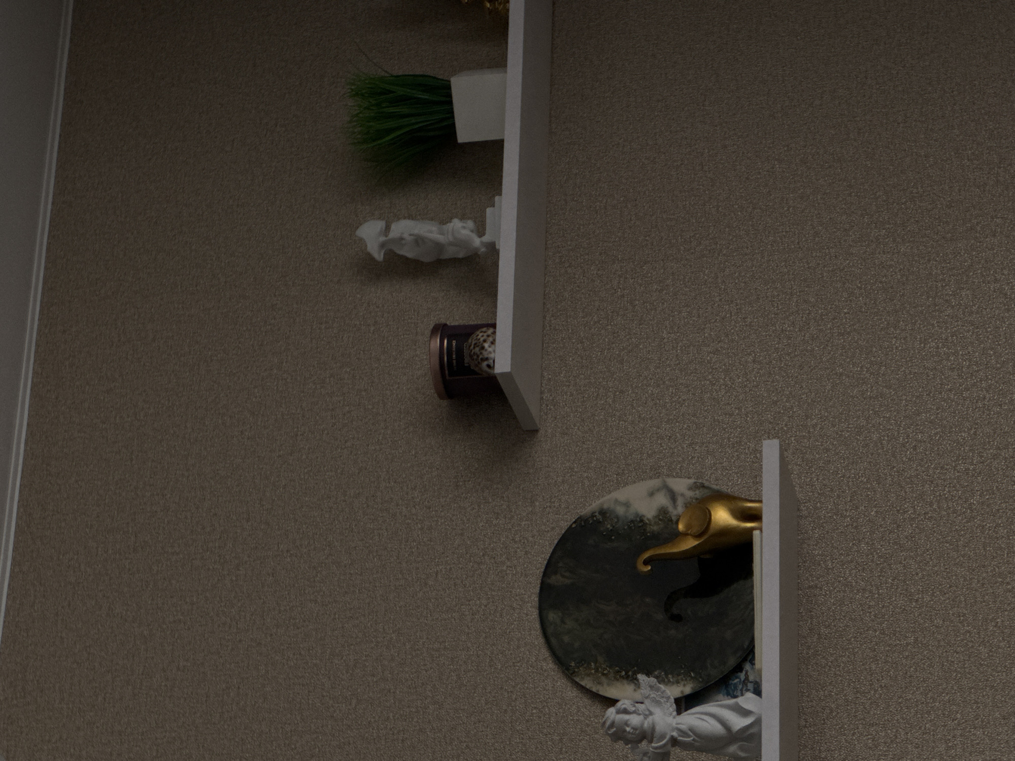 This media file captures an indoor scene in Chaikovsky, Russia, featuring wall-mounted decorative shelves against a textured, brownish-grey wall. No people are present, and no specific activity is taking place; the image is a static display of household decor.
On the upper visible white shelf, an artificial green grass or plant is displayed in a small white rectangular container. Adjacent to it lies a small, irregularly shaped white object, possibly a crumpled piece of paper or a small decorative item.
Below this, a dark, cylindrical object, resembling a candle or jar, is visible. It has a patterned label with some barely discernible text, appearing to start with "DONG..." or "DOVER..." and possibly including "COLLECTION." This object rests either on the lower part of the top shelf or on a separate, slightly lower shelf that is mostly obscured.
Further down, a white shelf supports a prominent round decorative disc. This disc features a dark, possibly marbled or resin-like surface with lighter, swirl-like patterns, resembling an artistic coaster or plate. Resting on this disc is a golden, abstractly shaped object with a distinct upward curve, which could be a stylized handle, a decorative element, or part of a small sculpture. Partially visible beneath this shelf and disc is a white sculpture, depicting what appears to be a cherub or angelic figure with detailed wings and facial features.
On the far left, a white vertical trim or doorframe is visible, indicating a structural element of the room. The lighting is dim and even, consistent with an indoor setting, and does not provide cues for the time of day.