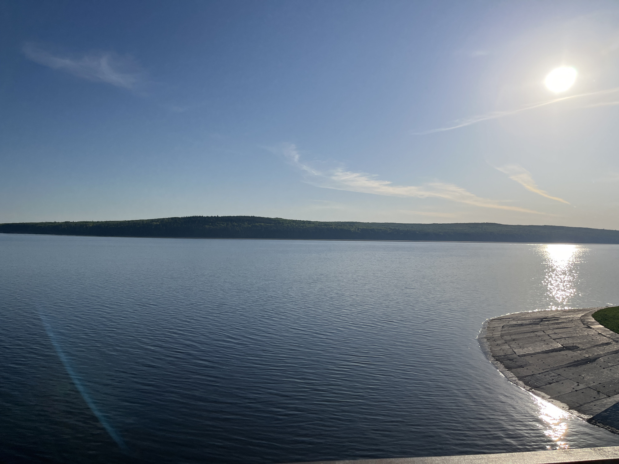 The image shows a tranquil view of the Kama River in Chaikovsky, Russia, under a clear blue sky with scattered clouds and the sun shining brightly. A long, wooded island stretches across the horizon, dividing the sky from the water. The river's surface is slightly rippled, reflecting sunlight with a bright glare on the right side of the image. A concrete structure, possibly a dam or embankment, is visible on the right edge of the frame, where the water meets the land. No people are visible.