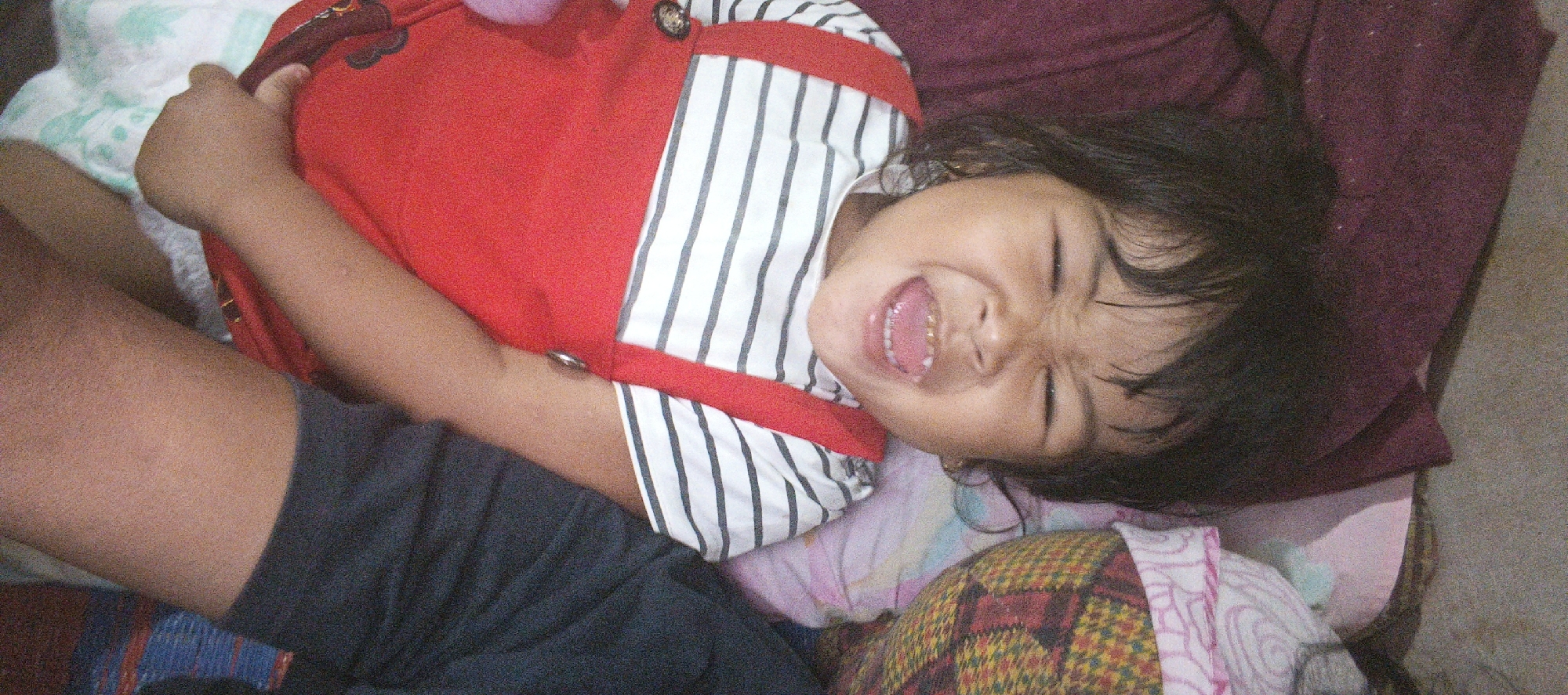 The image shows a close-up, overhead view of a young child lying down, possibly in a bed or on a soft surface. The child appears to be an infant or toddler with dark hair and is wearing a red garment with white and black striped suspenders. The child's mouth is open, revealing teeth, and their eyes are squinted, suggesting laughter, excitement, or a strong emotion. The child's left arm is visible, bent and resting on their chest. The background consists of patterned fabrics and a maroon-colored cloth, hinting at a domestic setting. The lighting suggests it could be daytime, but no specific time of day can be determined. There is no visible text in the image. The context of Cinta Dame, Indonesia, suggests this is a personal or family photograph taken in that region.