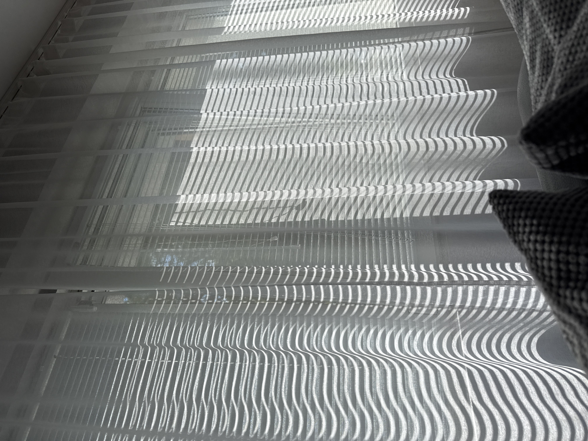 The image shows a close-up view of white sheer curtains with horizontal slats, causing a pattern of alternating light and shadow. A dark, textured object, possibly a pillow, is visible in the upper right corner, partially obscuring the view. The light suggests it is daytime. There is no information that can be attributed to the city of Lovech, Bulgaria.