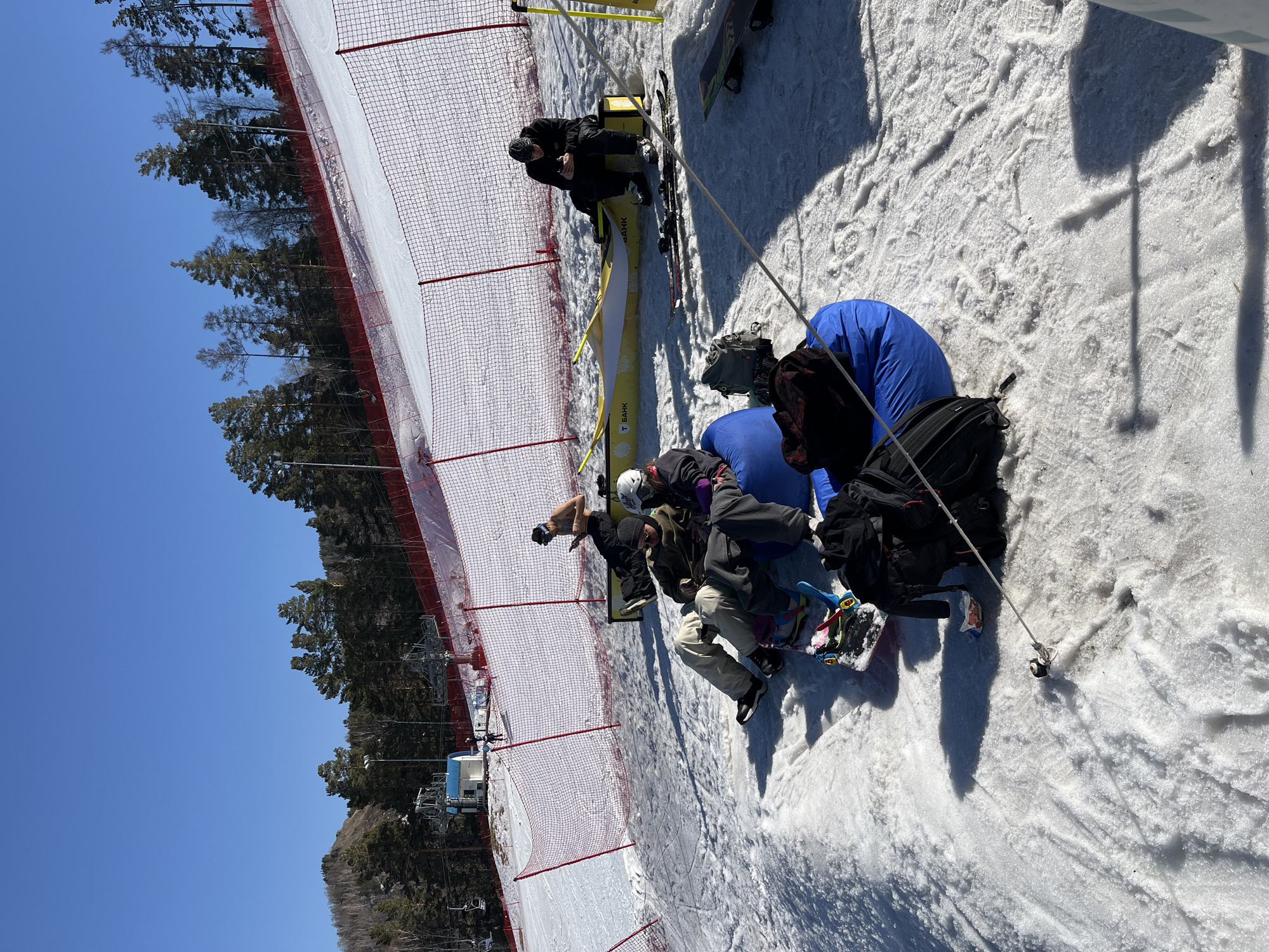 Several individuals are on a snow-covered slope near a red safety fence. They appear to be preparing for or resting after skiing or snowboarding, with skis and snowboards visible nearby. Some are sitting on blue pads. Trees line the area behind the fence, and a ski lift structure is also present. The sky is clear and blue. There is a yellow banner next to the people.
Location: Unknown, Unknown.