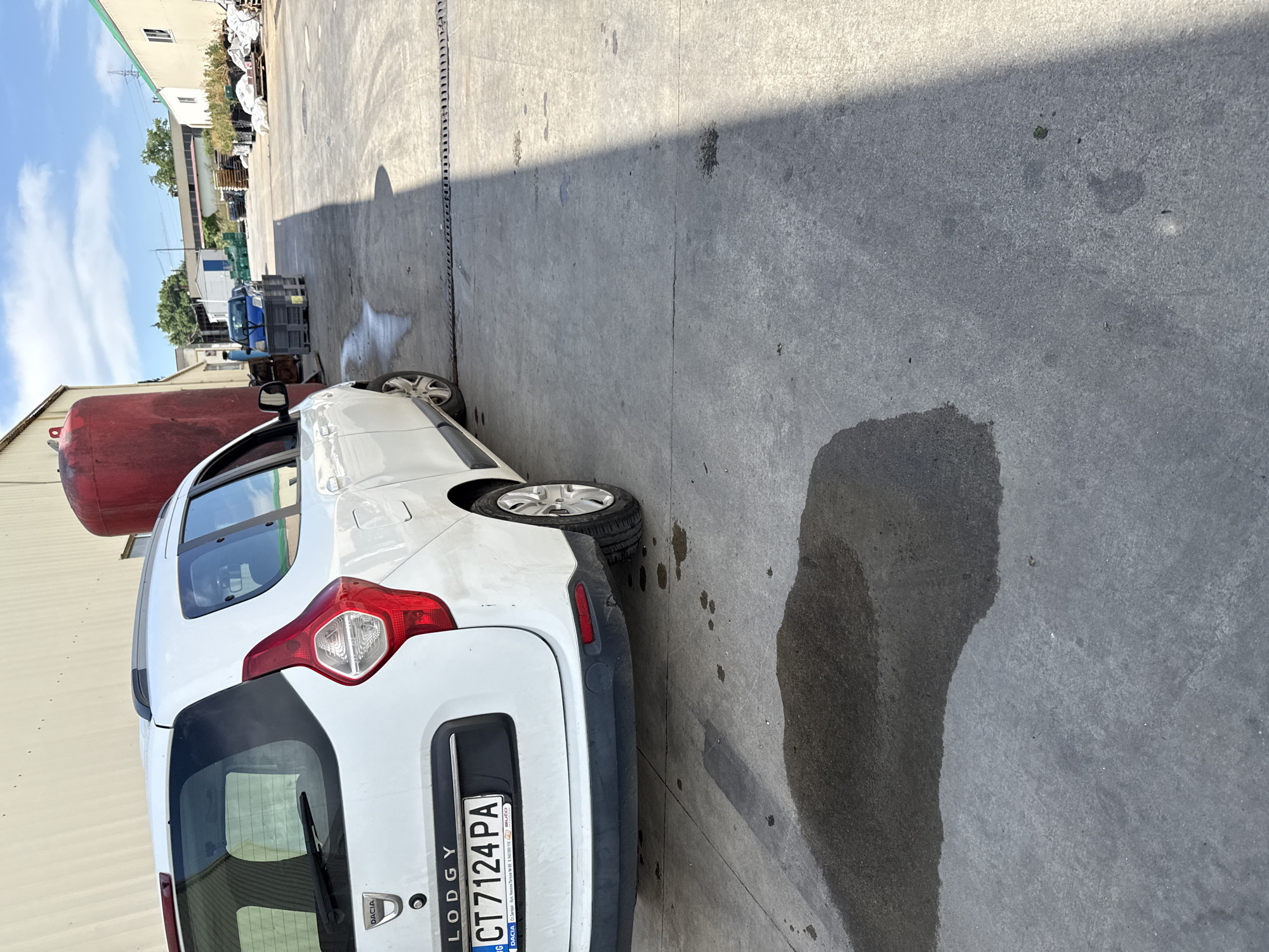 An eye-level outdoor shot captures the rear-left side of a white Dacia Lodgy car parked on a concrete surface under bright daylight with strong shadows. The car is positioned at an angle, facing away from the viewer towards a blue vehicle and buildings in the background. A large red industrial tank or container is visible to the left of the car. The concrete ground features several wet spots or puddles, including a significant dark patch behind the car and smaller drips near the rear tire. The sky is clear, suggesting sunny weather. The setting appears to be an industrial or commercial yard.
Notable details on the car include its model name "LODGY" and brand "DACIA" visible on the tailgate. The Bulgarian license plate reads "CT 7124 PA," indicating the car is registered in Stara Zagora. Below the license plate, smaller text from a dealer sticker is partially visible, stating "DACIA Ситра буд. Никола Петков № 69" along with a phone number. The car's rear bumper shows some minor dirt or scuff marks. A metal chain runs along the edge of the concrete surface on the left.