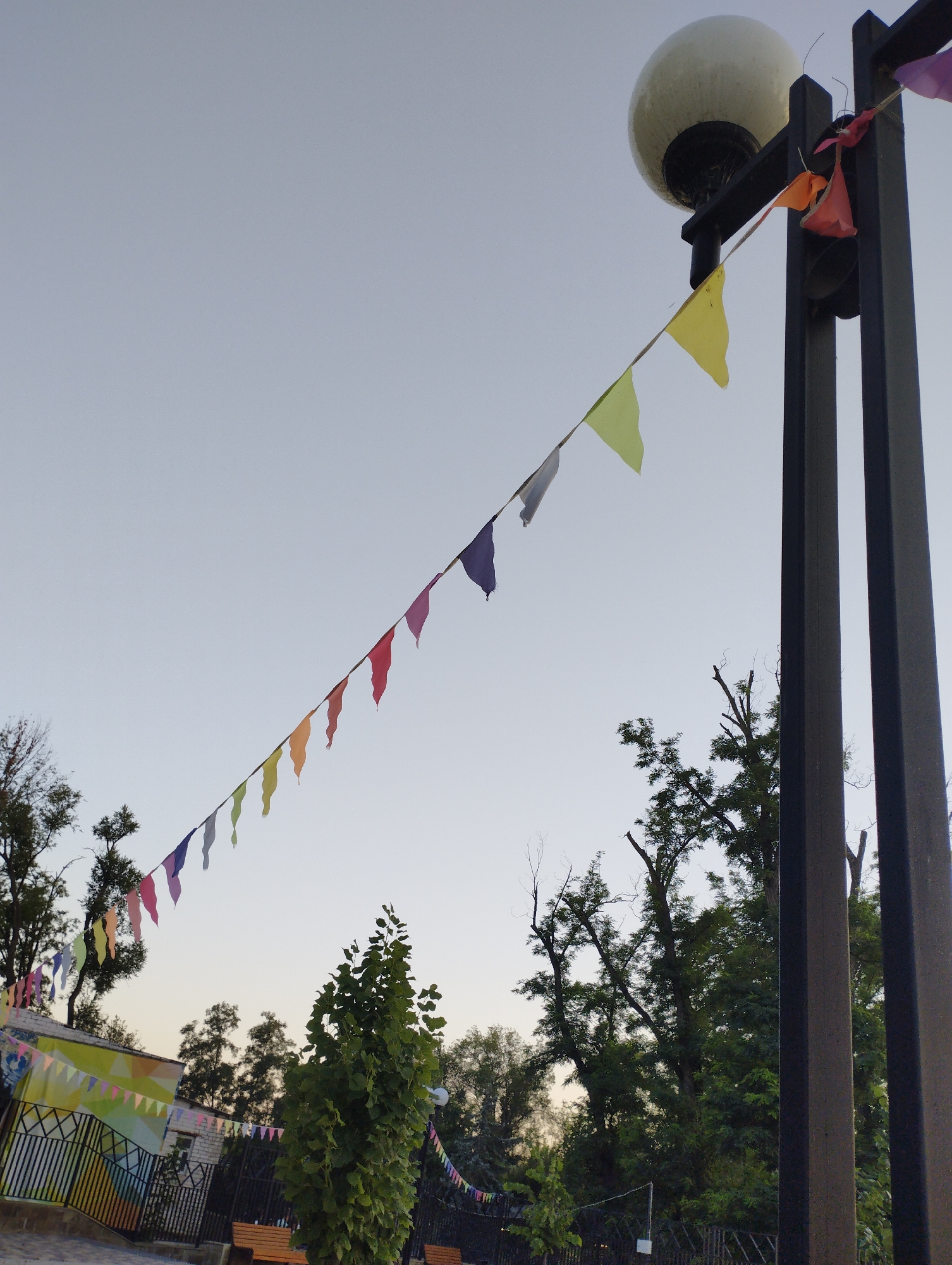 The image shows a street lamp with a globe-shaped light fixture, decorated with a string of small, colorful triangular flags. The flags are strung across the sky, from the lamp post to an unseen point off-camera. In the background, there are trees, a fence, and a building with a colorful mural. The scene appears to be an outdoor public space, possibly a park or a street in a town. There are no people visible in the image. The sky is a pale blue, indicating daytime, possibly early morning or late afternoon given the soft lighting. The flags are likely a festive decoration, suggesting an event or celebration. The overall impression is one of a quiet, possibly slightly overgrown, outdoor area.