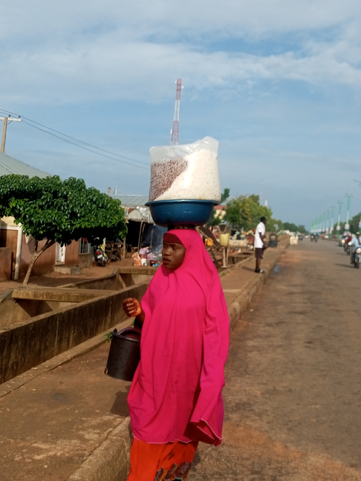 A woman in a pink hijab and a pink abaya is walking on a sidewalk in Tumfure, Nigeria. She is carrying a large bowl of rice on her head, and a black bucket is hanging from her left arm. The sidewalk is made of concrete and is lined with a low concrete wall. In the background, there is a street with traffic, including motorcycles and cars. There are also several market stalls and trees. The sky is blue with some clouds.