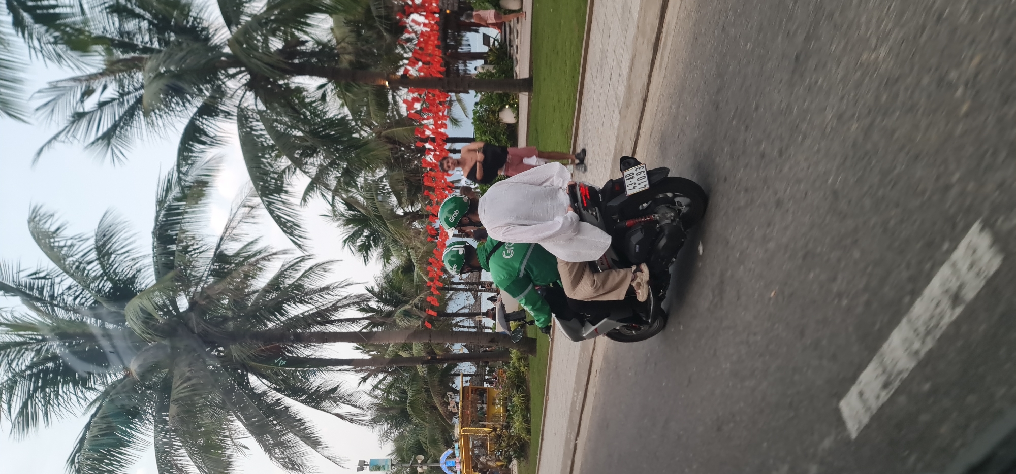 The image depicts an outdoor scene in Đà Nẵng, Vietnam, likely during the late afternoon or early evening. The foreground features a paved road with visible white lane markings. On this road, a black motorcycle is seen carrying two individuals. The driver is wearing a green uniform and matching helmet with the prominent "Grab" logo, indicating a ride-hailing service. The passenger, seated behind the driver, wears a white long-sleeved shirt and light-colored pants. The motorcycle's license plate, "43-B 410.93," is clearly visible, with "43-B" being a common identifier for vehicles registered in Đà Nẵng province.
To the left of the road, a wide paved sidewalk or pathway runs parallel, separated by a curb. Further left, there's a grassy area densely populated with numerous tall palm trees, suggesting a tropical or coastal environment. Between some of these palm trees, strings of vibrant red decorations, possibly small lanterns or flags, are hung, hinting at a festive or special occasion. Several other people are visible in the background, some walking on the sidewalk and one person standing near the red decorations, observing the surroundings. The sky appears bright and clear.