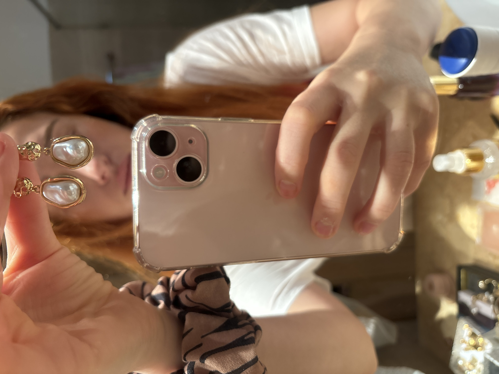 A selfie, potentially in Budva, Montenegro given the prompt's context, shows a fair-skinned person with red hair holding an iPhone. The phone is centered, slightly angled, with a clear case. A gold earring with a white pearl hangs near the left edge of the frame. The person wears a white t-shirt. A hair tie with a brown and black animal print pattern is visible. The background appears to be a table with cosmetics and jewelry, reflected in a mirror-like surface. One white and gold cosmetic bottle is partially visible in the upper right. The overall lighting is bright and natural.
